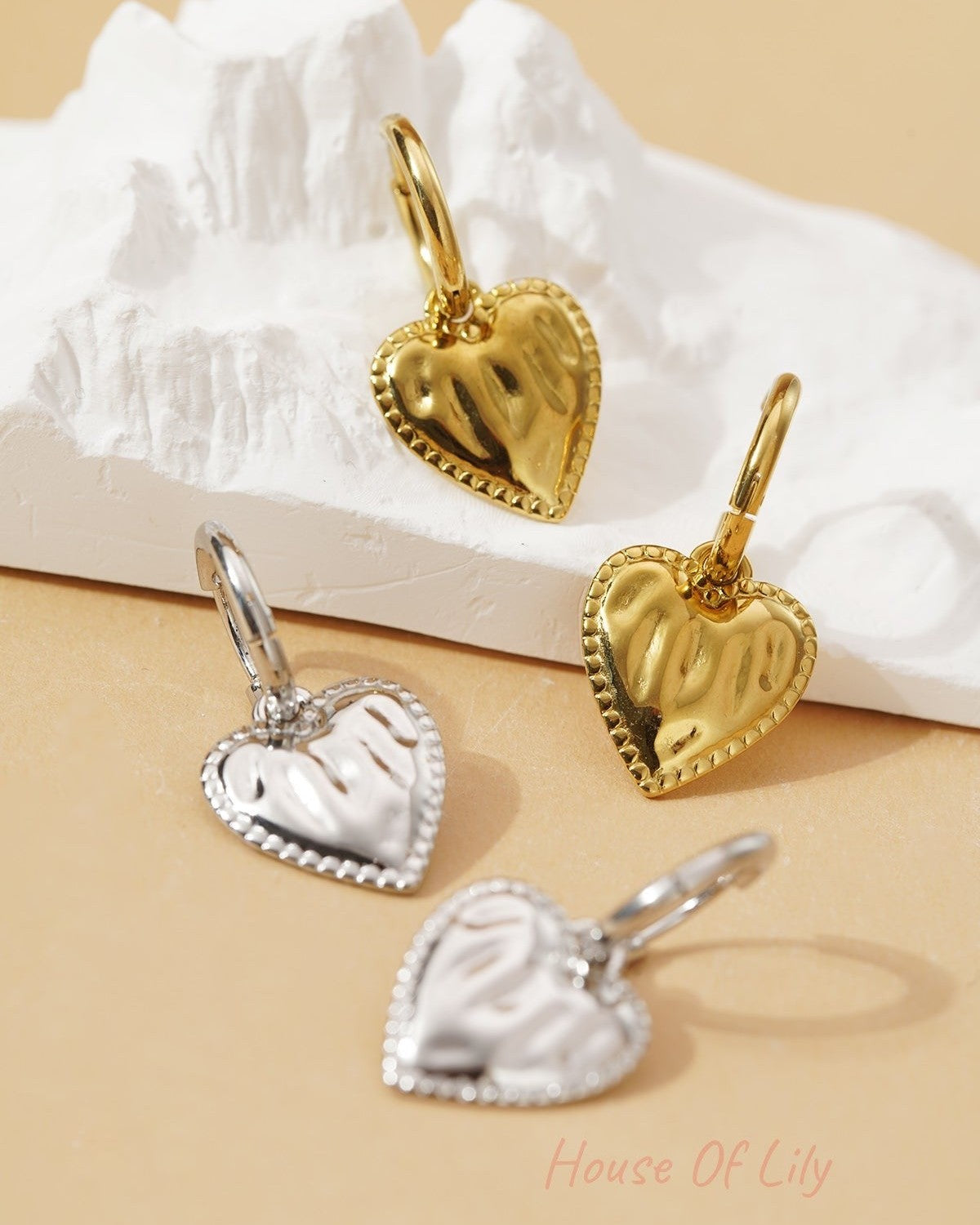 Gold and silver Sculpted Coquette heart charm hoop earrings with a beaded border, resting on a white textured base.