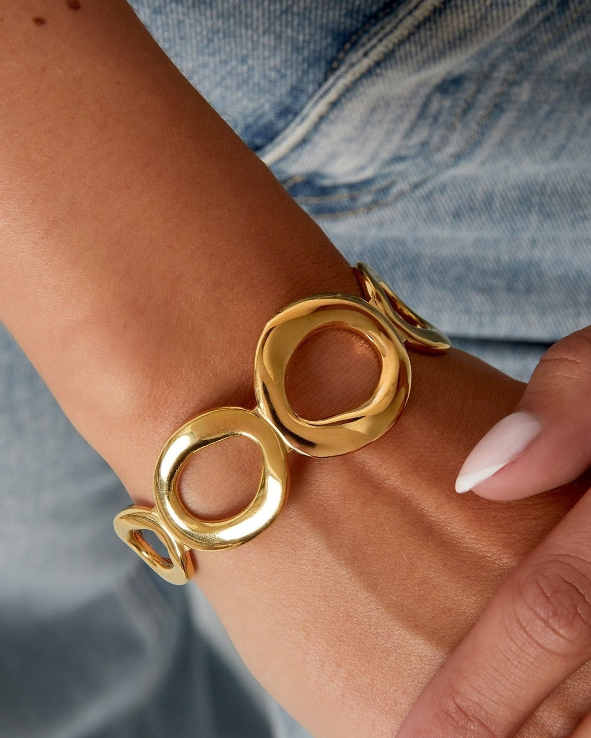 Close-up of a woman's wrist wearing a shiny gold openwork cuff bracelet featuring irregular open oval shapes paired with denim outfit