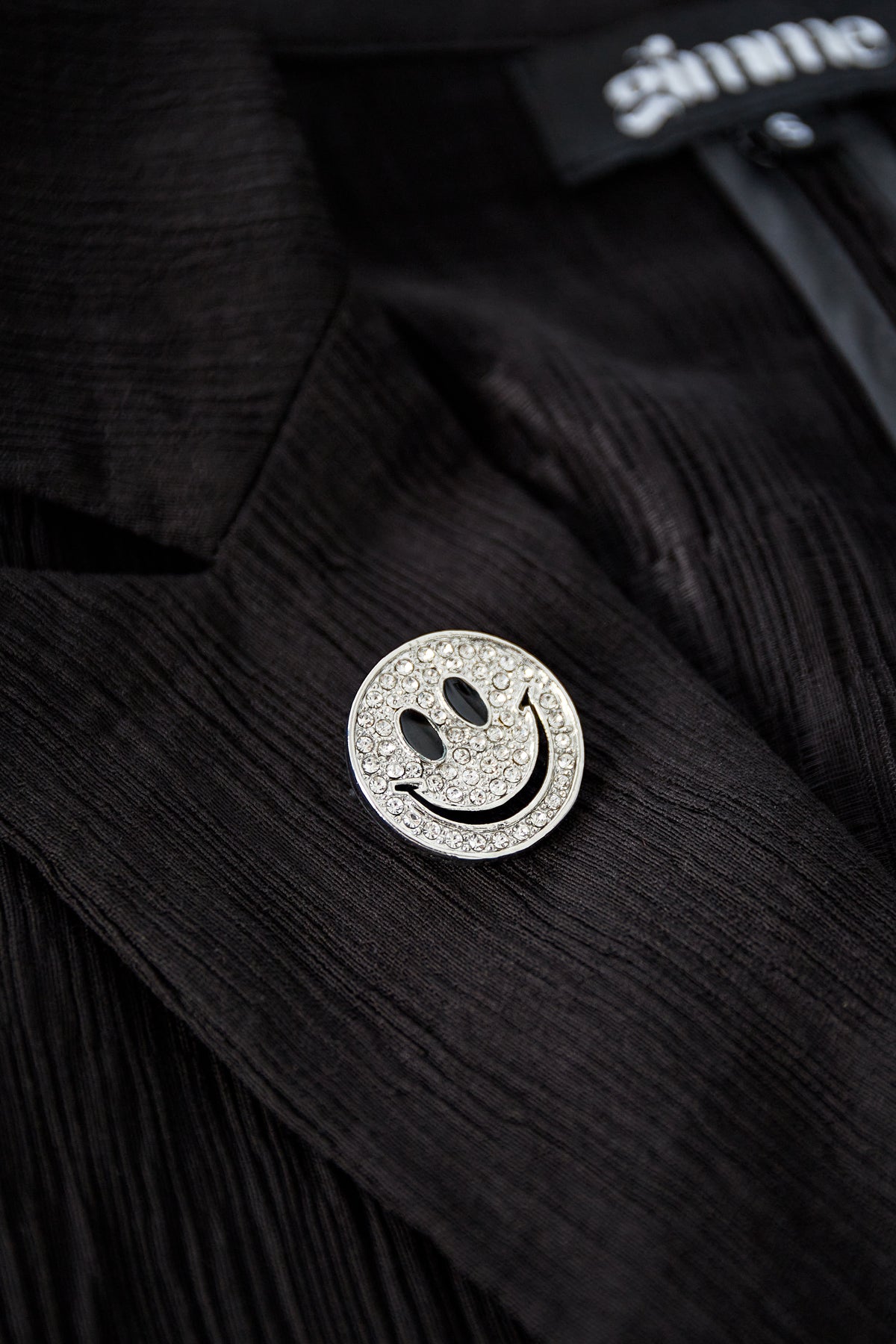 Silver-tone smiley face brooch with clear crystals pinned to a black textured jacket