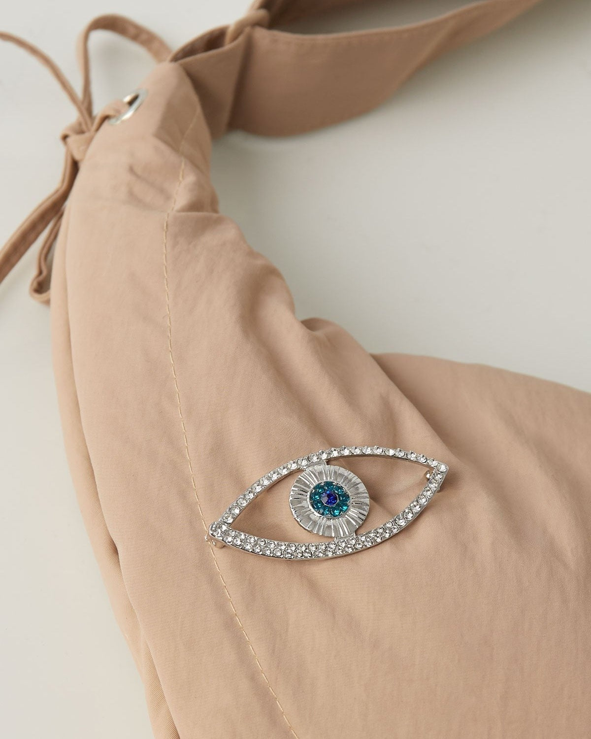 Silver-tone evil eye brooch pinned on a beige fabric bag with visible clear and blue crystals