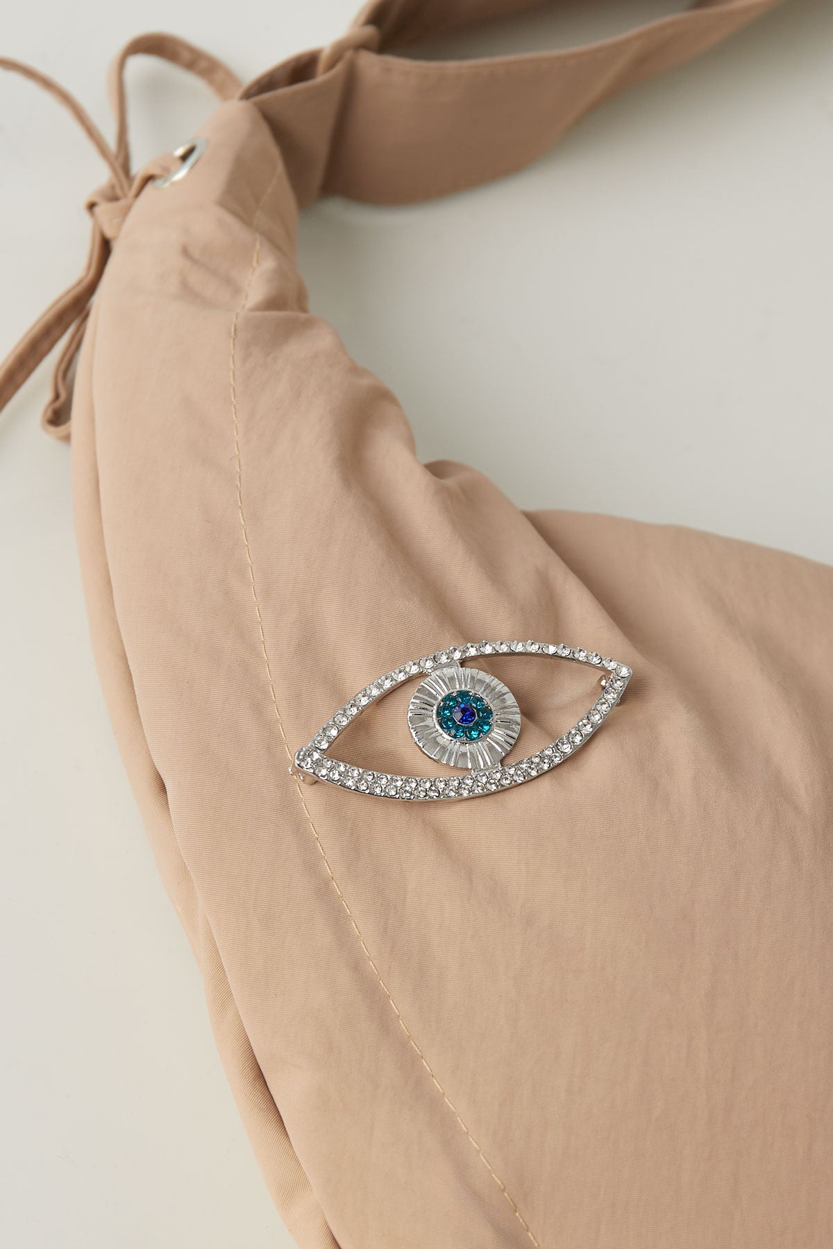 Silver-tone evil eye brooch pinned on a beige fabric bag with visible clear and blue crystals