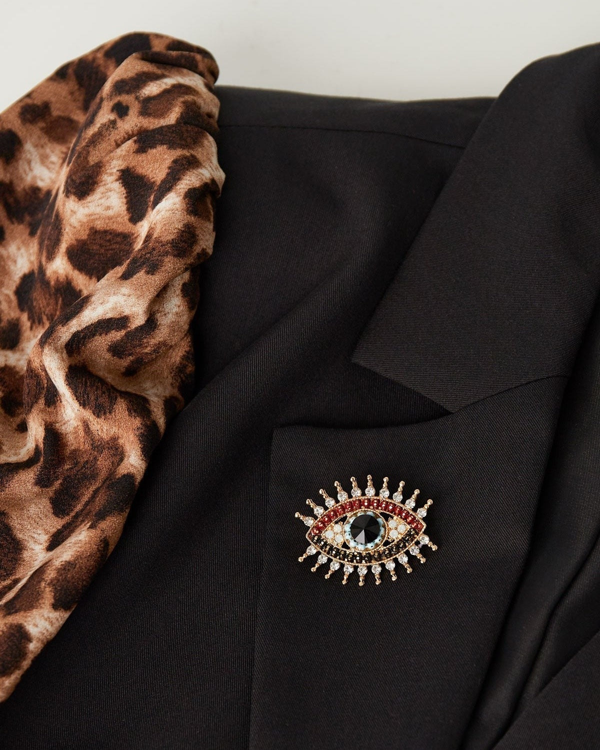 Charming eye brooch pinned on a black blazer next to a leopard print scarf, showcasing a stylish and youthful accessory look