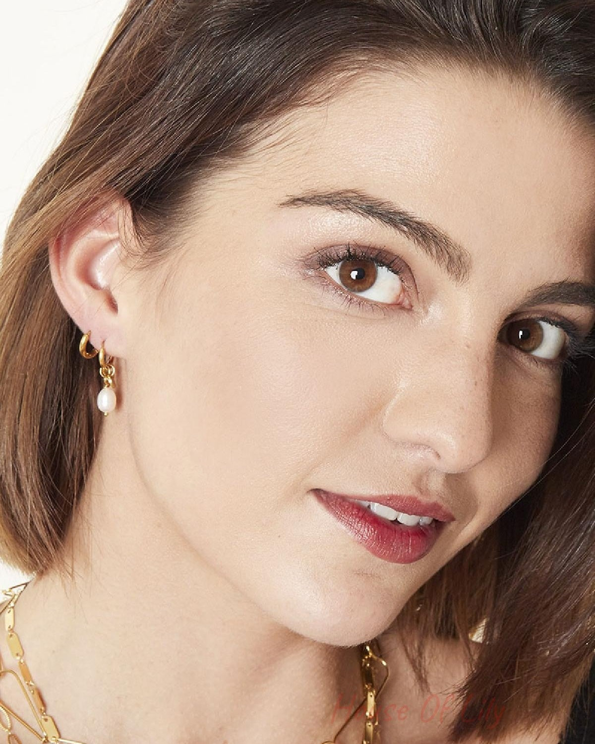 Close-up portrait of a smiling woman wearing gold pearl drop hoop earrings, styled with layered gold necklaces