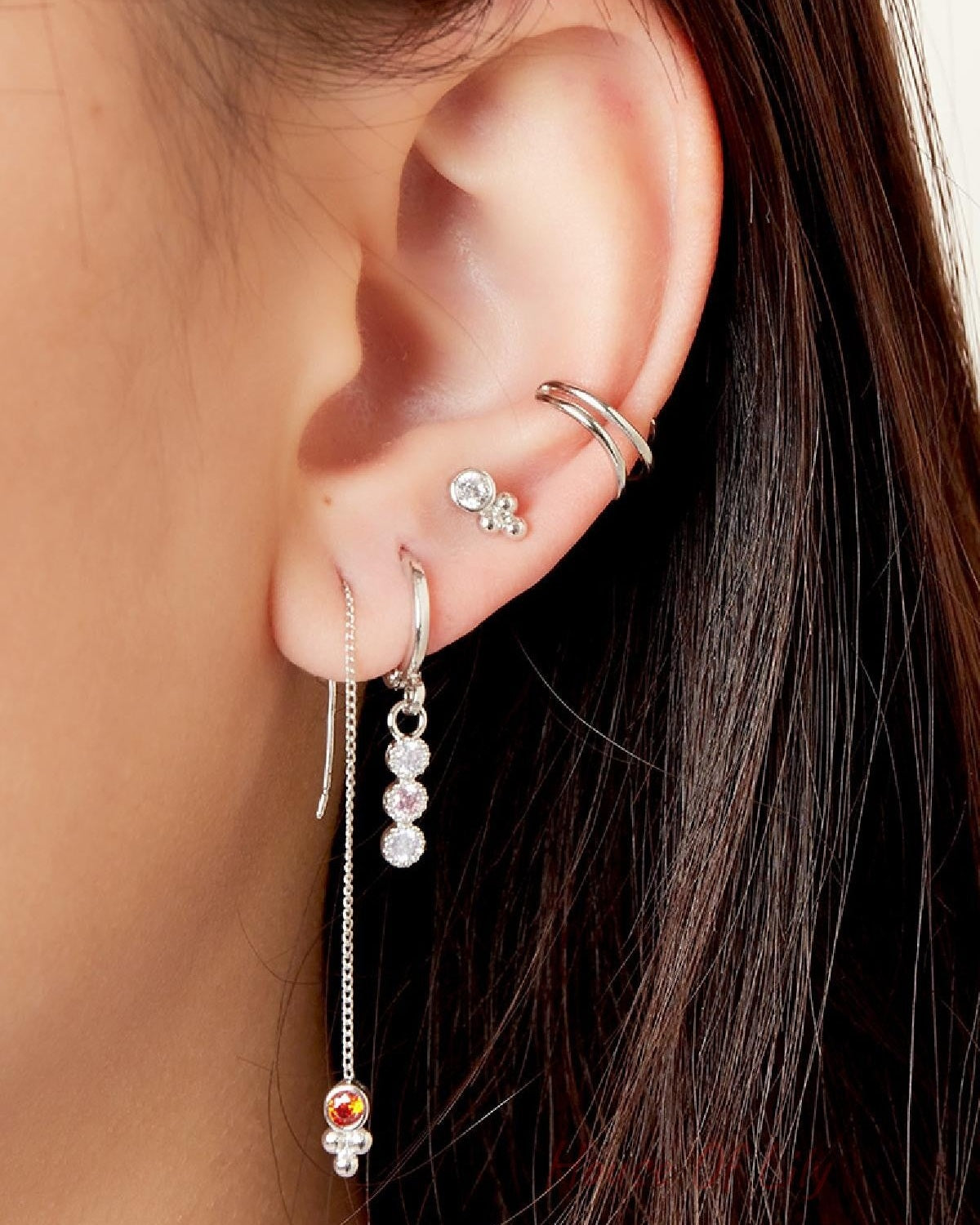 Close-up photo of an ear wearing silver earrings including a silver spiral ear cuff, dangling earrings with crystals, and small studs, showcasing elegant ear styling.