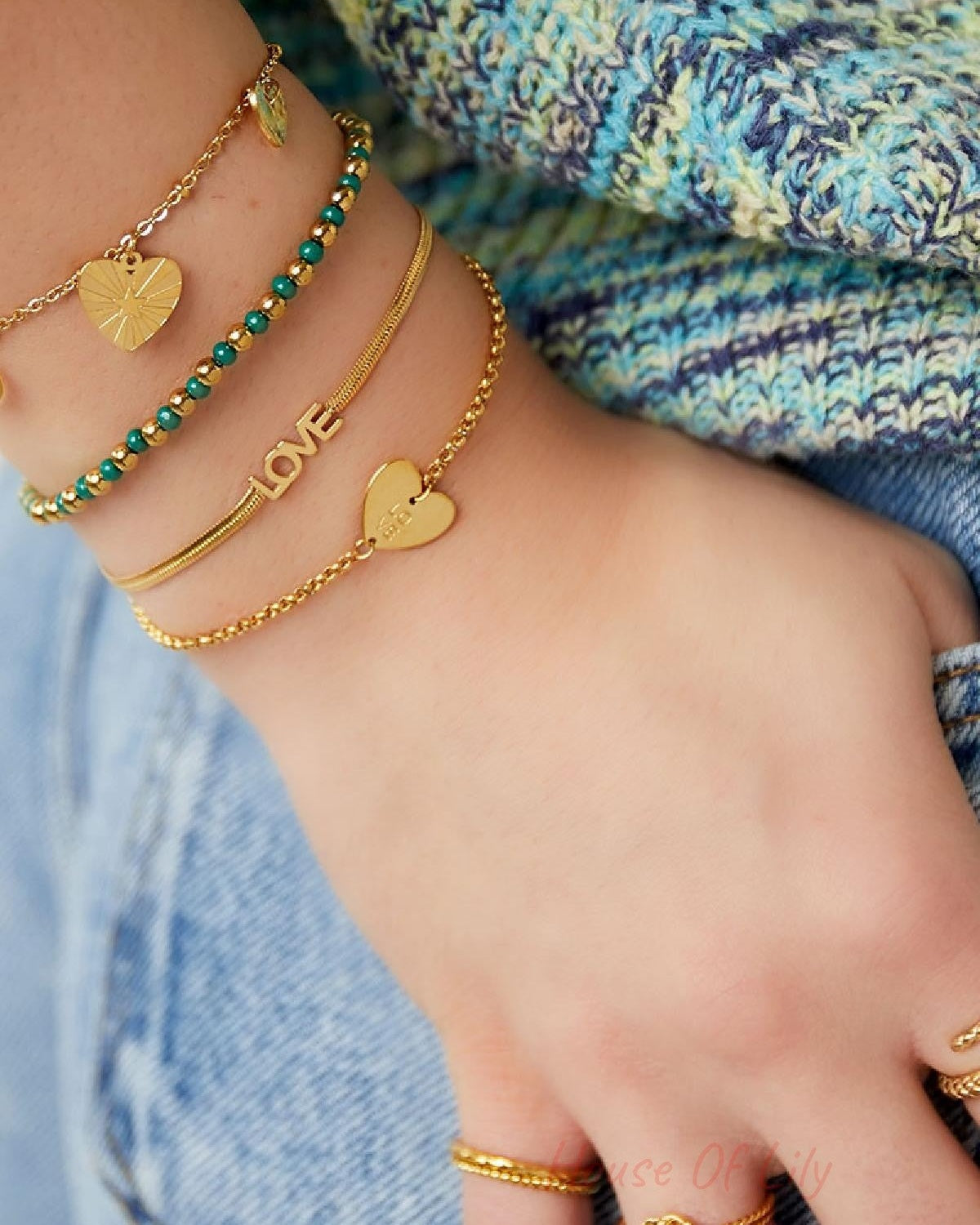 Close-up of a woman's wrist adorned with layered bracelets including the gold 'LOVE' charm bracelet, complemented by other gold and green bead bracelets, against a casual textured sweater and denim