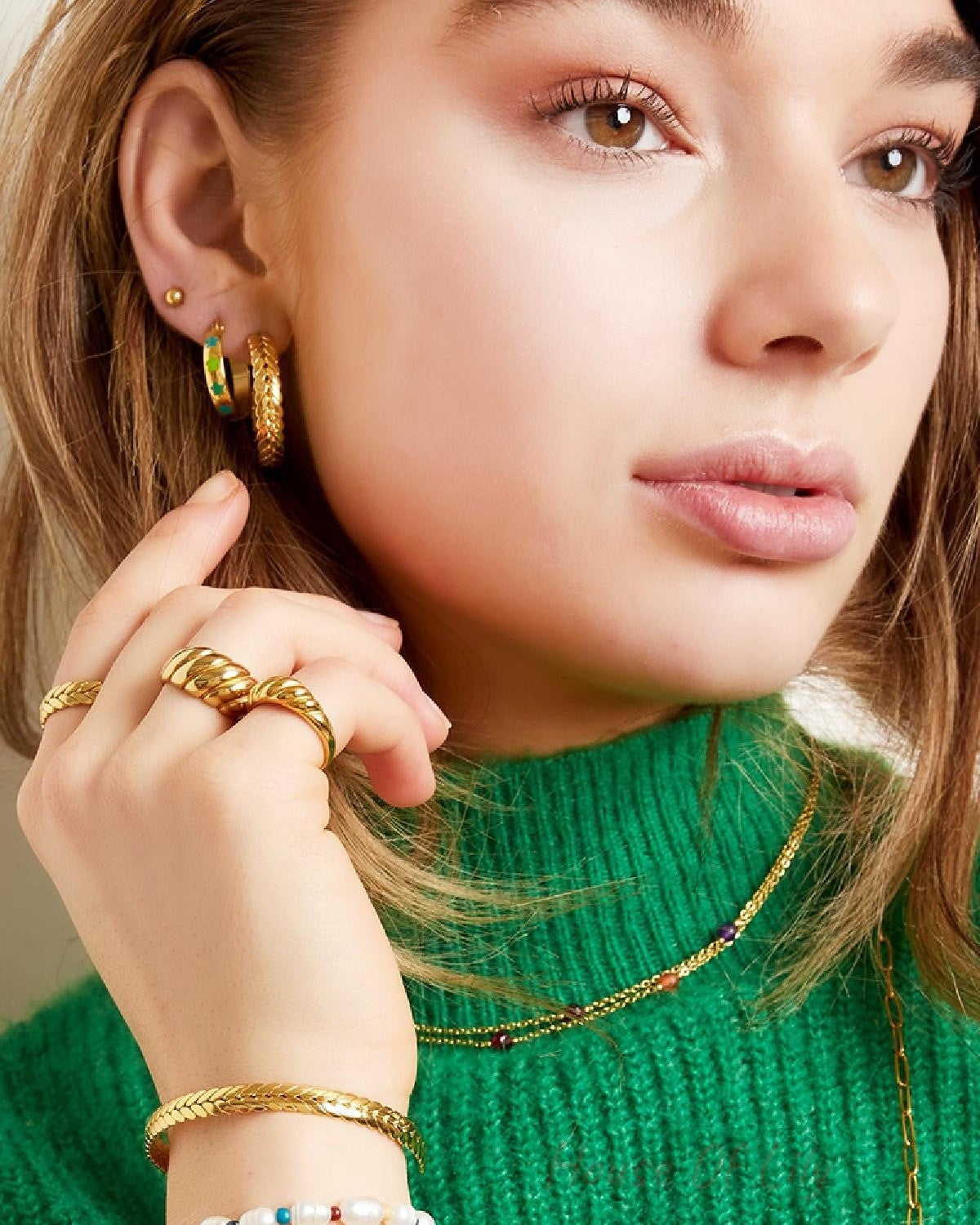Close-up of woman showing gold laurel cuff bracelet, rings, and layered necklaces on green sweater