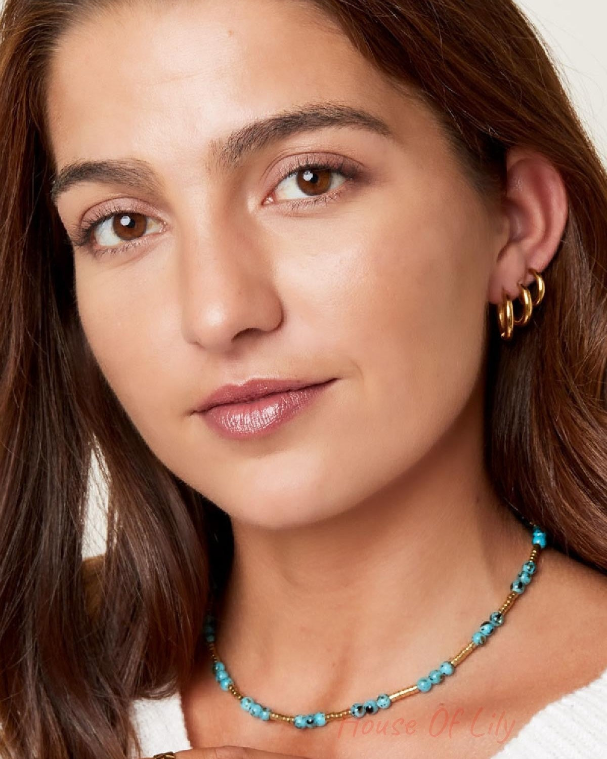 Front-facing portrait of a smiling woman wearing three gold hoop earrings and a turquoise beaded necklace