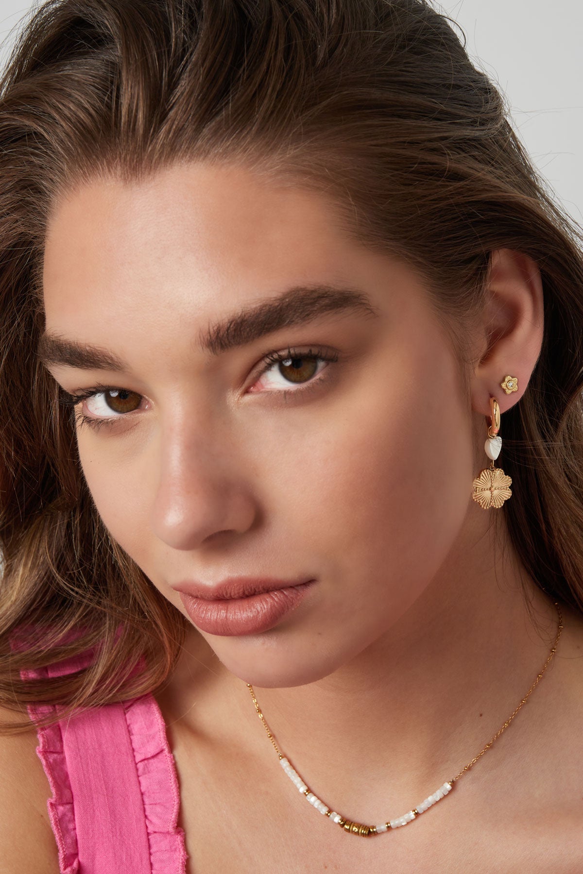 Model close-up showing youthful styling with gold flower stud earrings worn with a pink ruffled top and beaded necklace