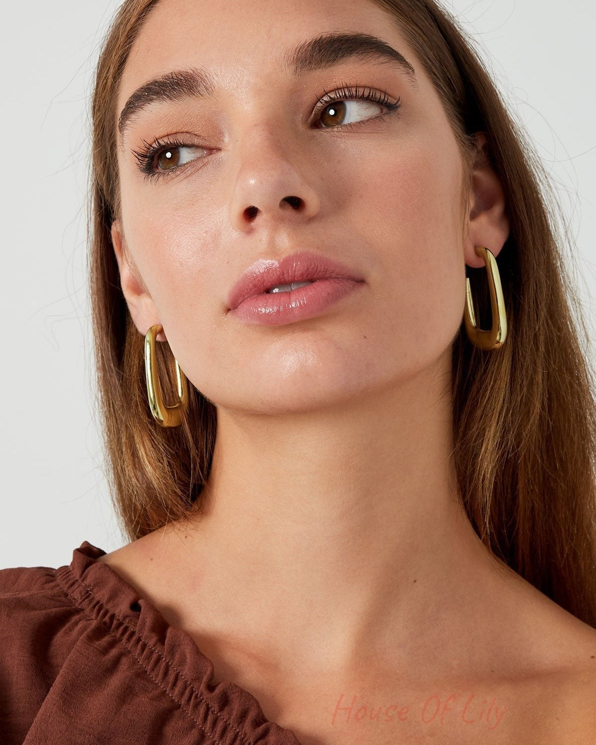 Close headshot of woman wearing polished gold rounded rectangle hoop earrings with subtle smile and natural hair