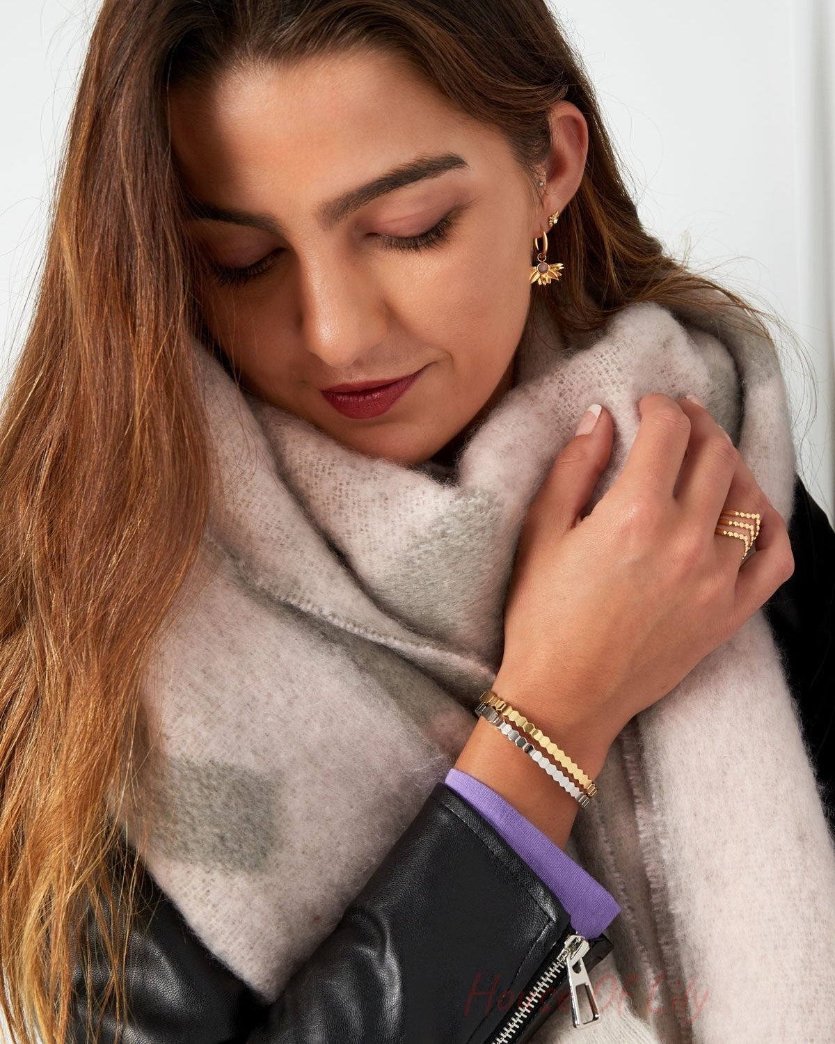 Woman showing wrist with stacked gold and silver hexagon link bracelets, beige scarf, and black jacket, head tilted down