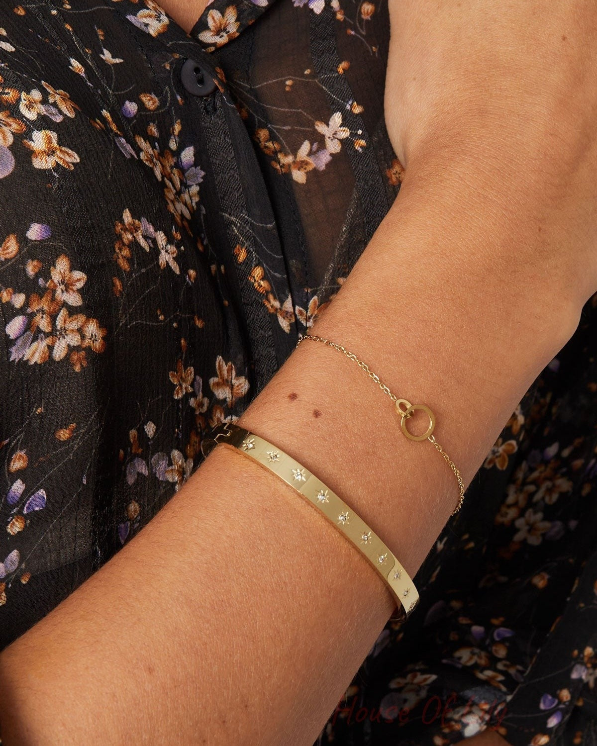 Close-up of a woman’s wrist wearing gold-tone star-studded bangle paired with a delicate gold chain bracelet and floral sheer blouse