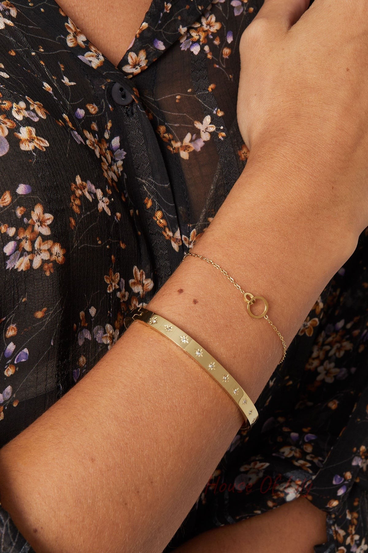 Close-up of a woman’s wrist wearing gold-tone star-studded bangle paired with a delicate gold chain bracelet and floral sheer blouse