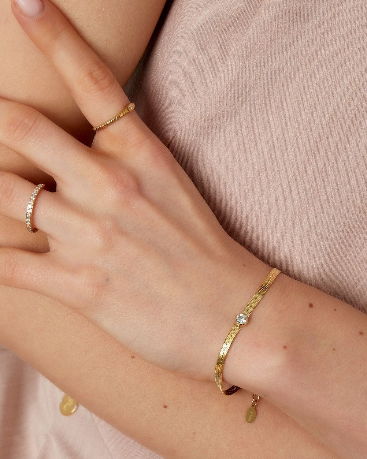 Woman's wrist wearing a slim gold chain bracelet with a sparkling crystal, shown with delicate gold rings against a soft beige top