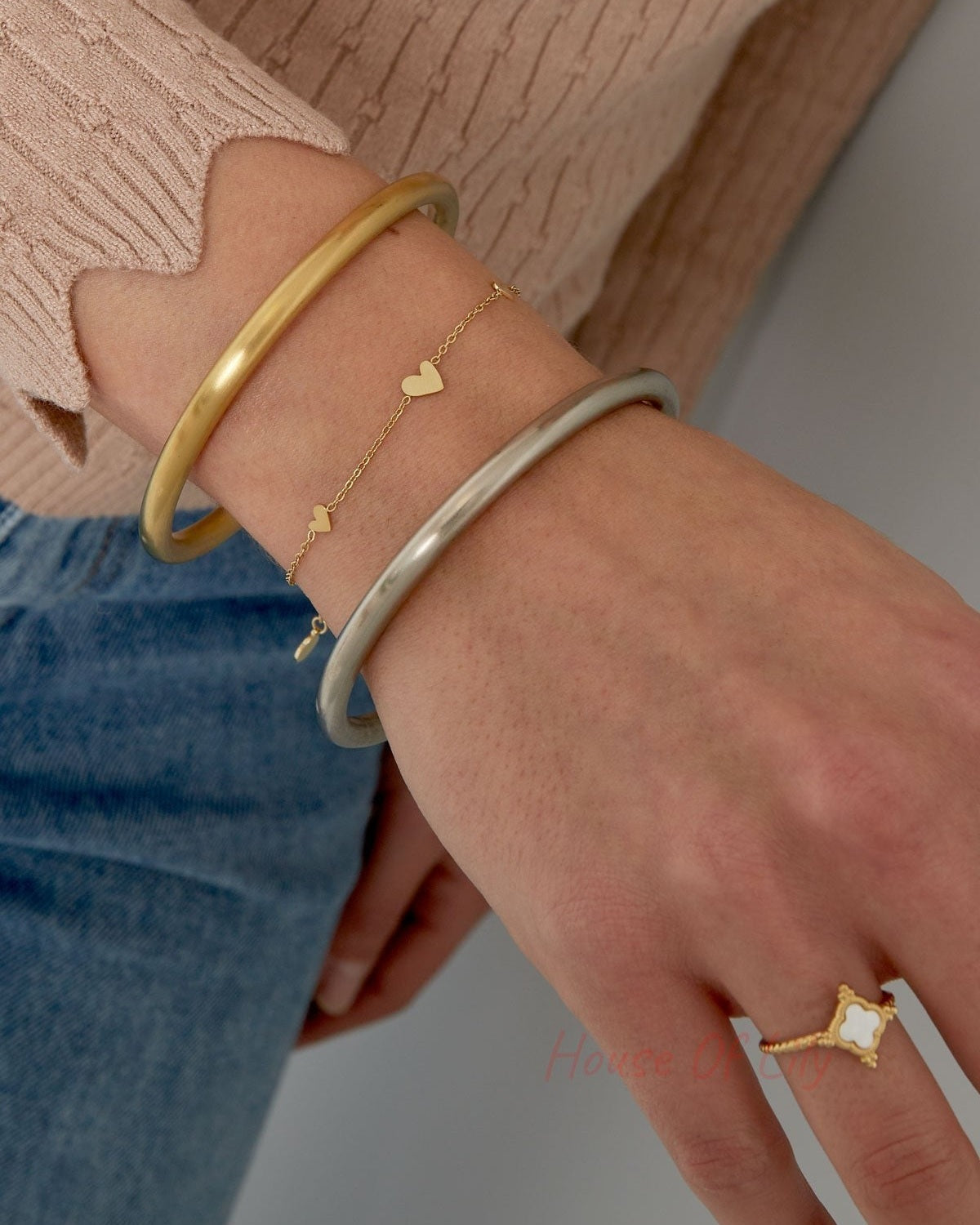 Close-up of a woman's wrist wearing the delicate gold heart charm bracelet alongside two bangles, styled with beige sweater and blue jeans