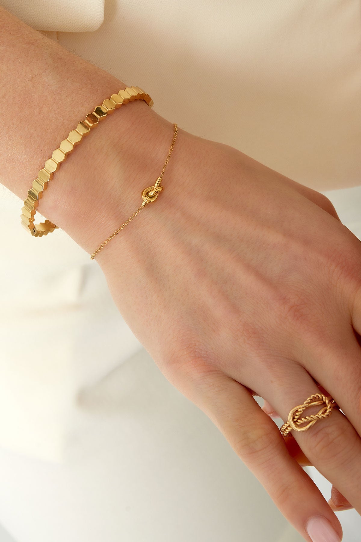 Close-up of a woman's wrist wearing a delicate gold knot bracelet paired with a hexagonal gold bangle and a twisted gold ring