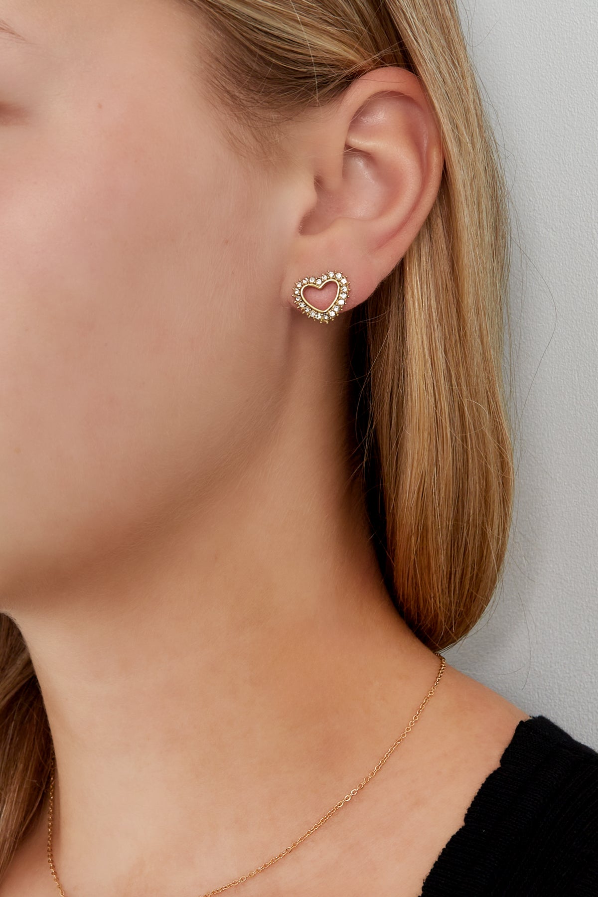 Woman wearing gold-tone rhinestone heart stud earrings with minimal makeup and black top