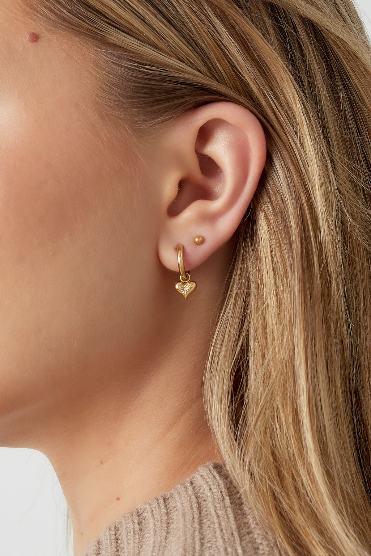Female model wearing a golden heart charm hoop earring paired with a small golden stud, showcasing the earring’s size and style