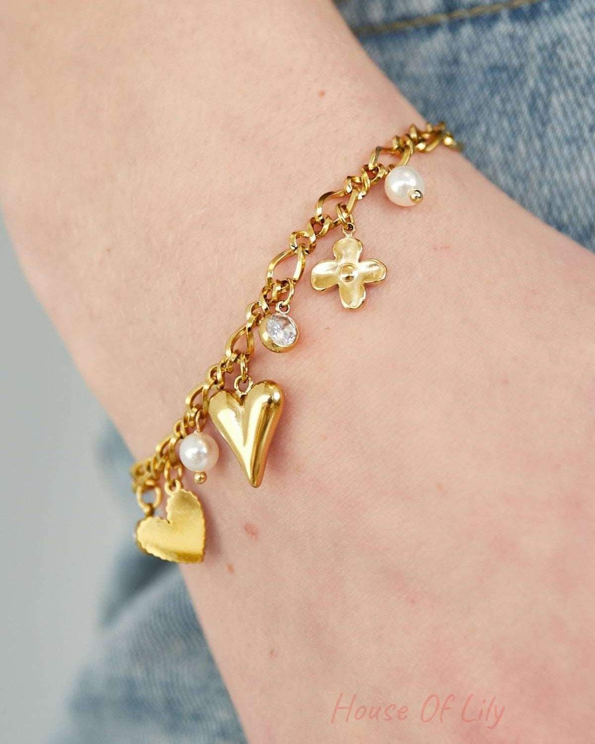 Close-up of the gold charm bracelet worn on wrist, showing detailed heart, flower, rhinestone and pearl charms paired with denim