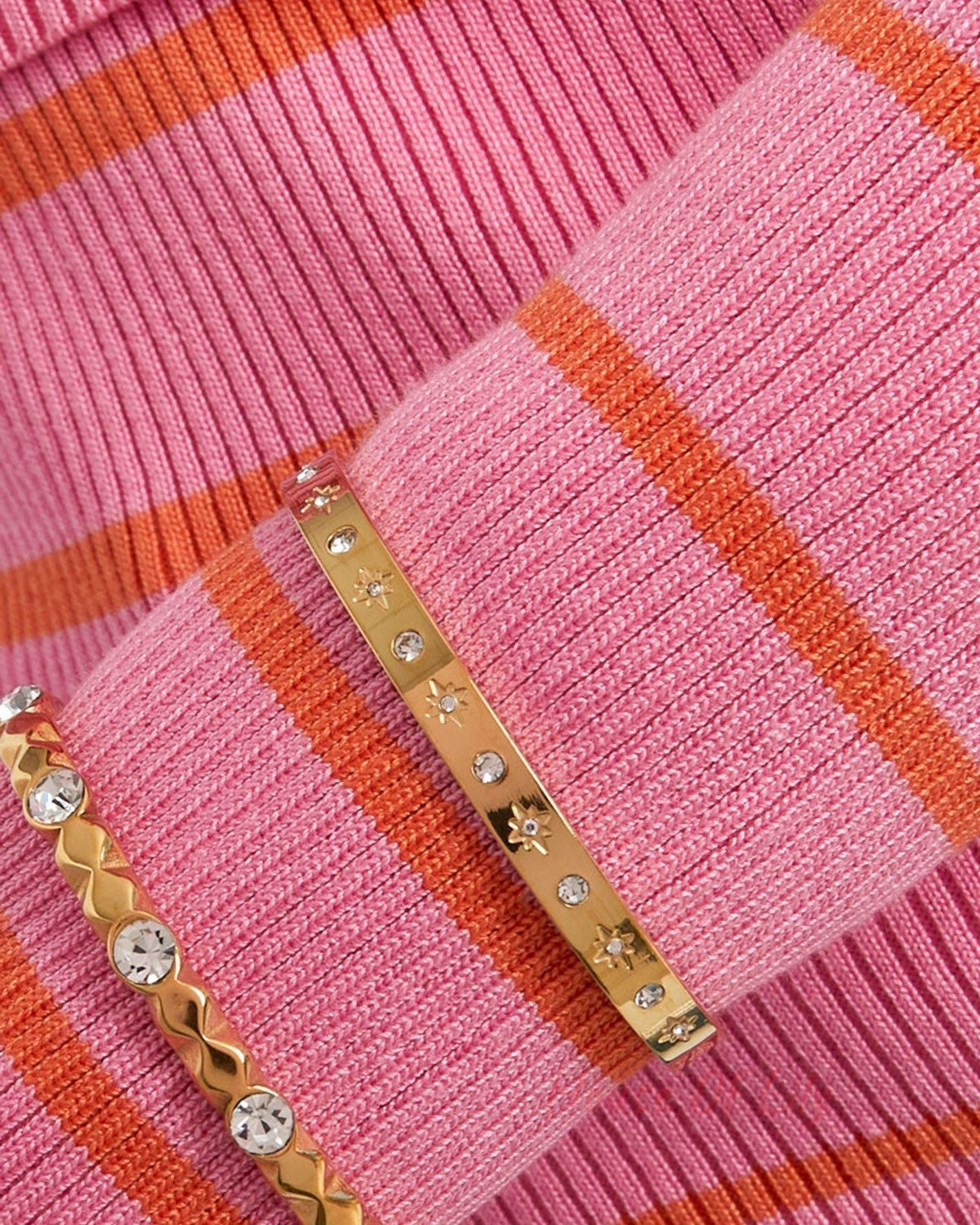 Close-up of a gold-tone starry bracelet worn over a pink and orange striped sweater, paired with another crystal bracelet