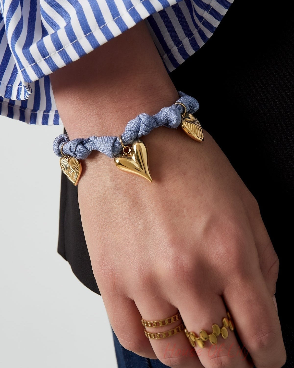 Model wearing blue braided heart charm bracelet with three gold heart pendants on wrist and gold rings