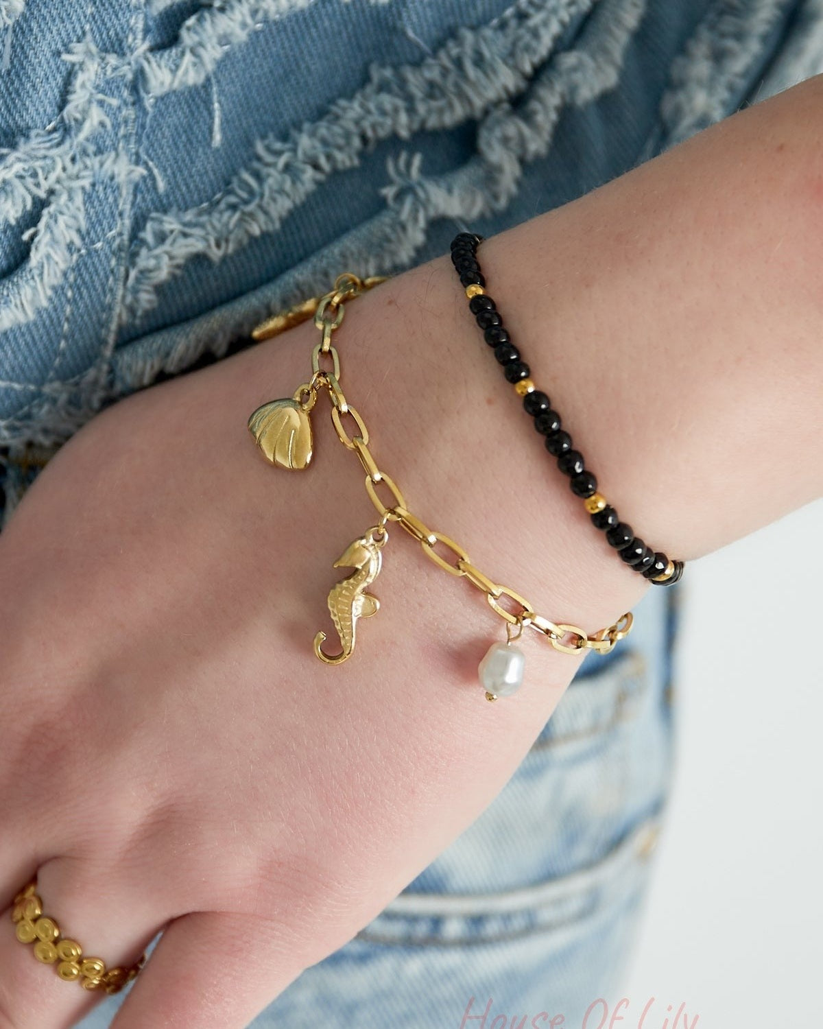 Close-up of wrist wearing a black and gold beaded bracelet paired with a gold charm bracelet featuring seashell, seahorse, and pearl charms over denim jeans