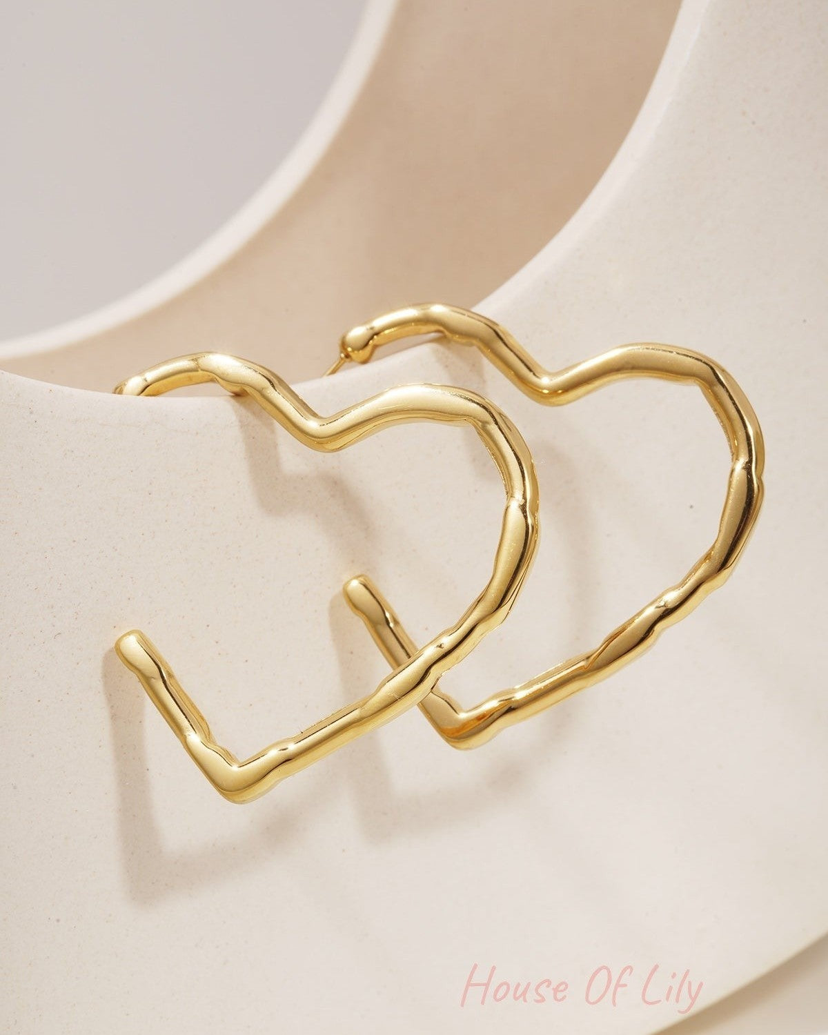 Gold-plated heart-shaped hoop earrings with a textured, organic finish, resting against a cream ceramic stand.