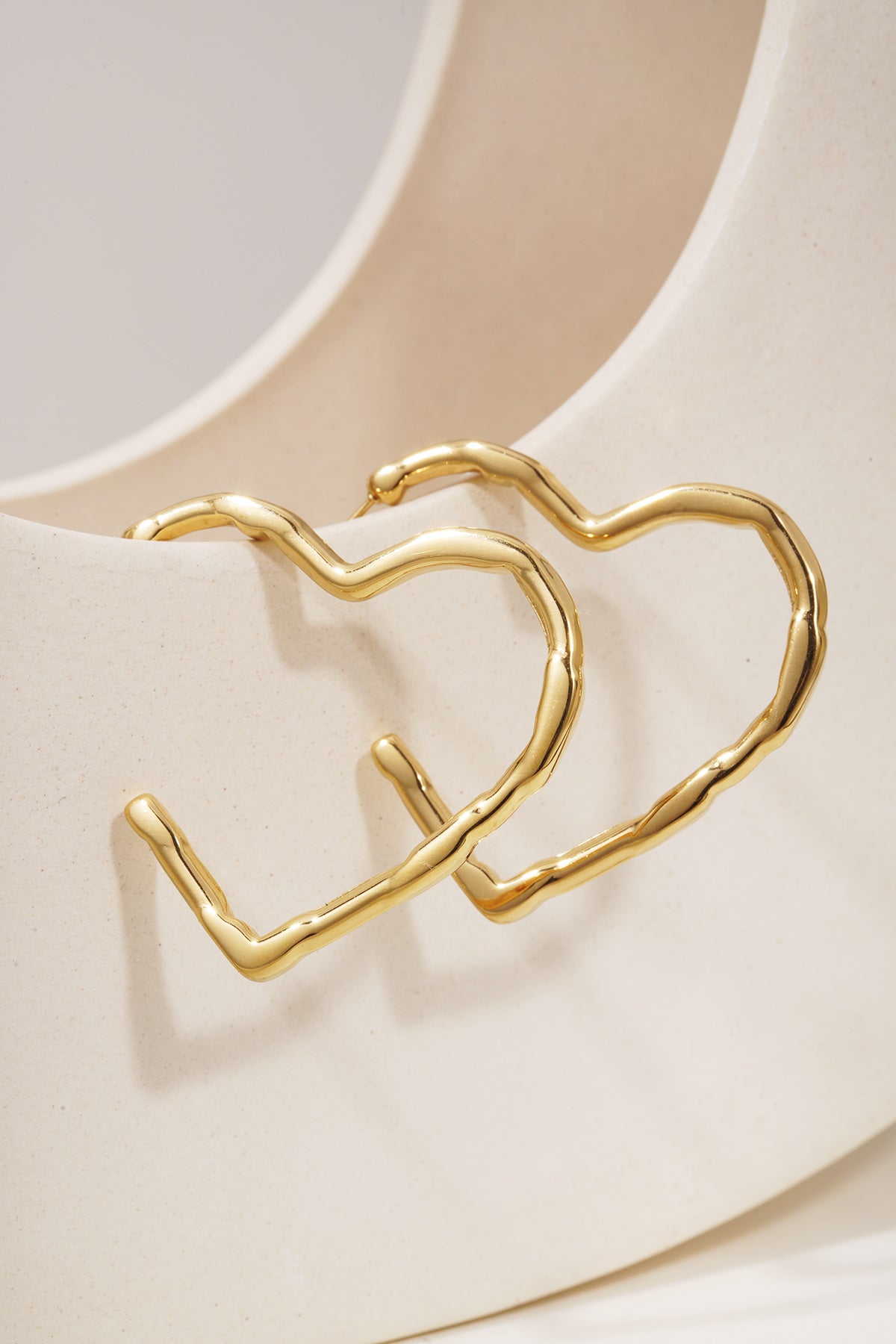 Gold-plated heart-shaped hoop earrings with a textured, organic finish, resting against a cream ceramic stand.