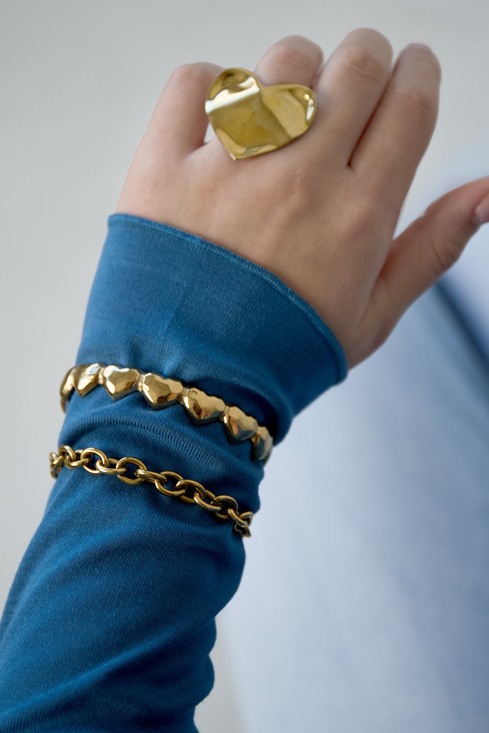 Gold Bold Link Chain Bracelet stacked with a heart-link bracelet, worn on a blue sleeve with a large gold heart ring.