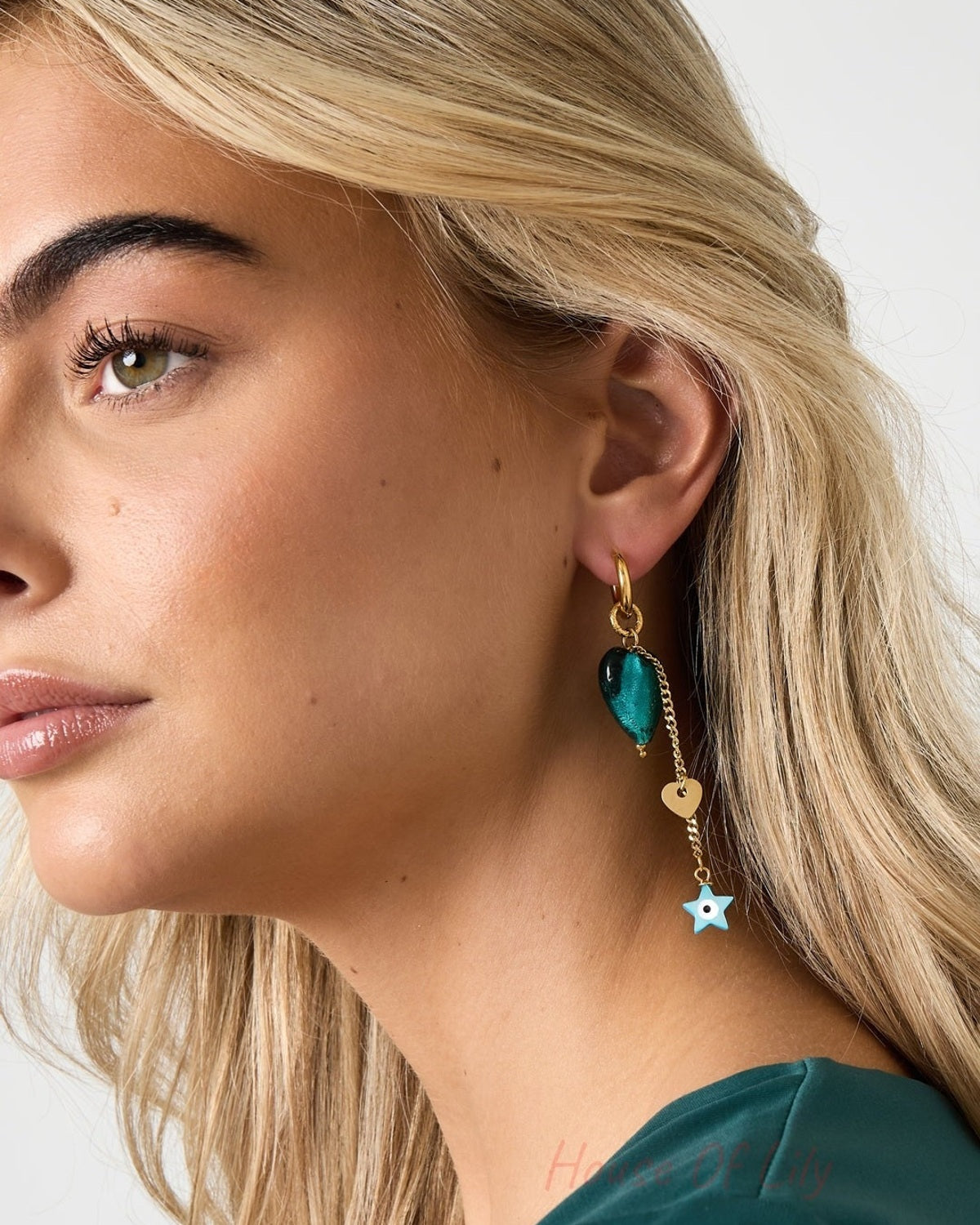Side view of a woman wearing gold-tone drop earring with emerald-green heart, small gold heart charm, and blue star pendant dangling
