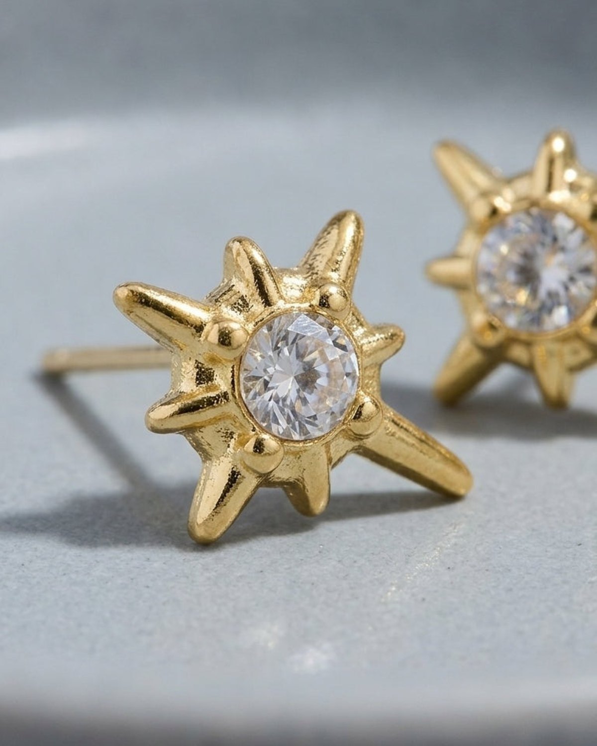 Gold starburst stud earrings featuring a central round-cut clear crystal, set in a textured, polished gold finish.
