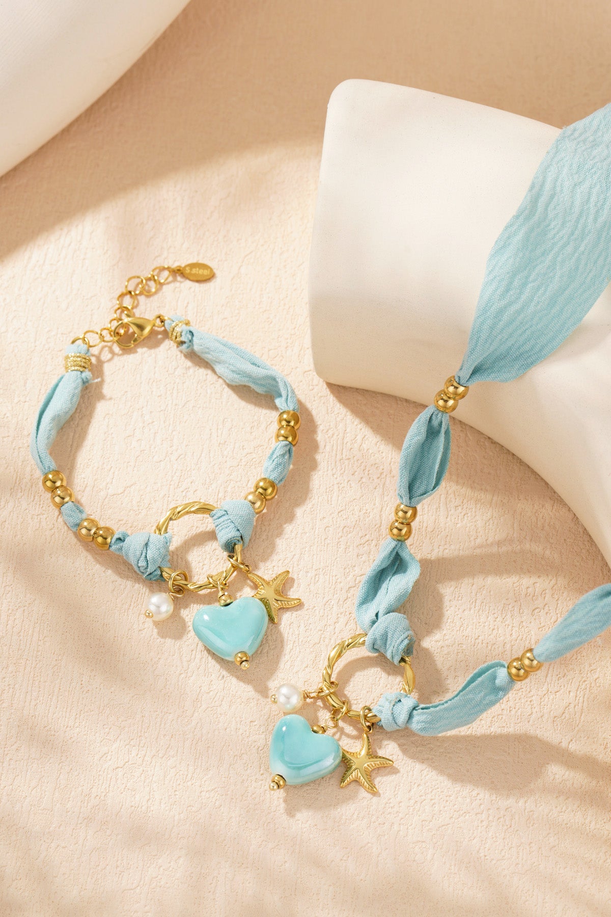 Matching braided heart charm necklace and bracelet set laid on textured cream surface, highlighting gold beads and pastel charms.