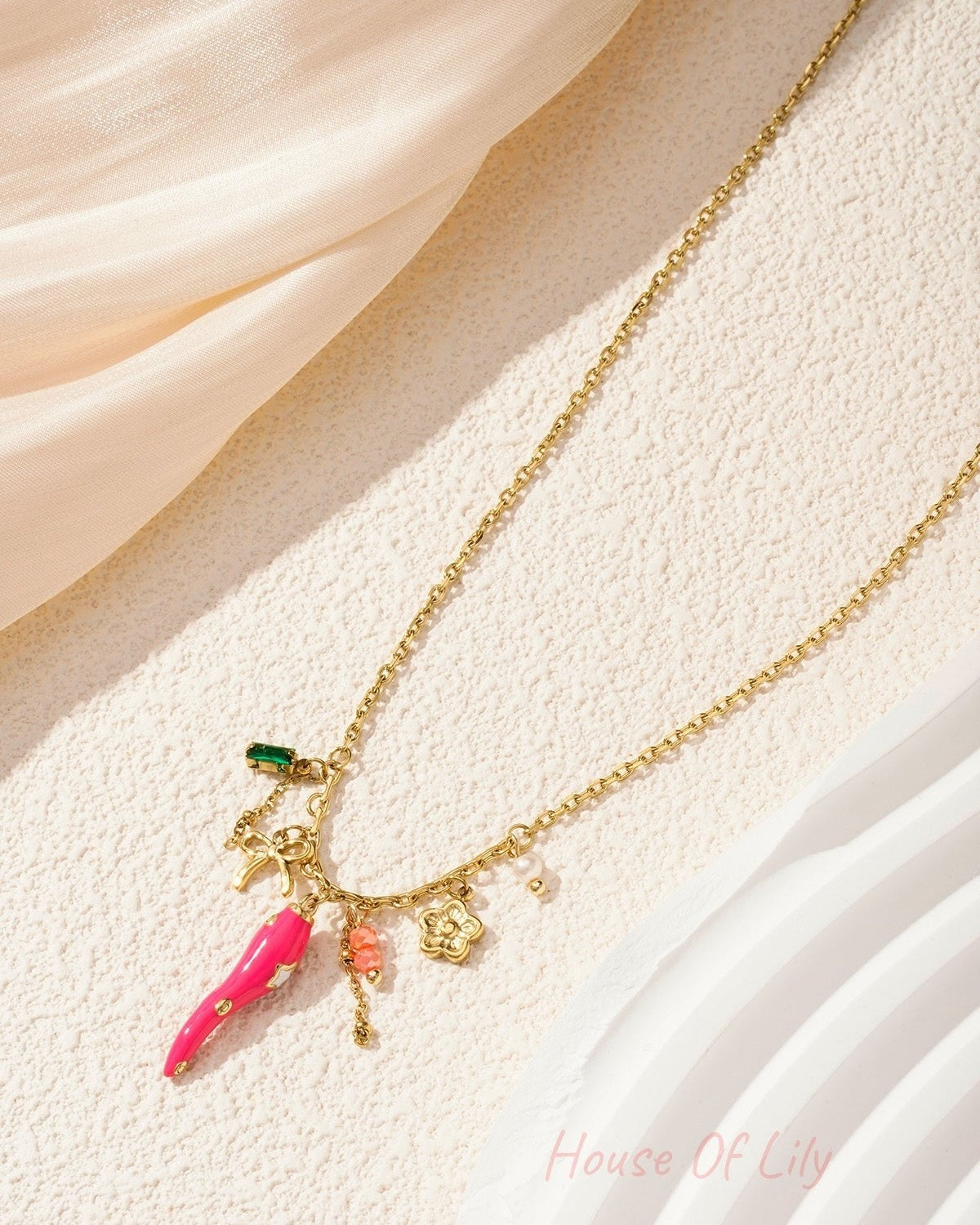Gold-tone pendant necklace with vibrant pink pepper charm and floral accents styled on neutral fabric and stone background