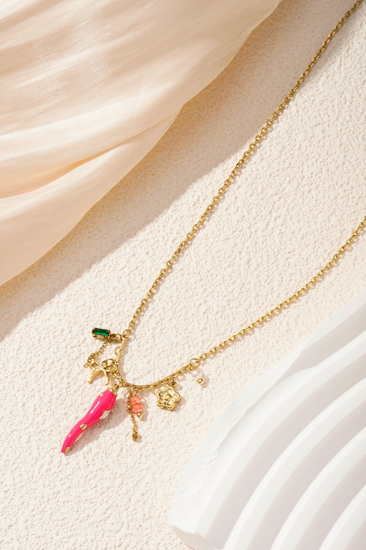 Gold-tone pendant necklace with vibrant pink pepper charm and floral accents styled on neutral fabric and stone background