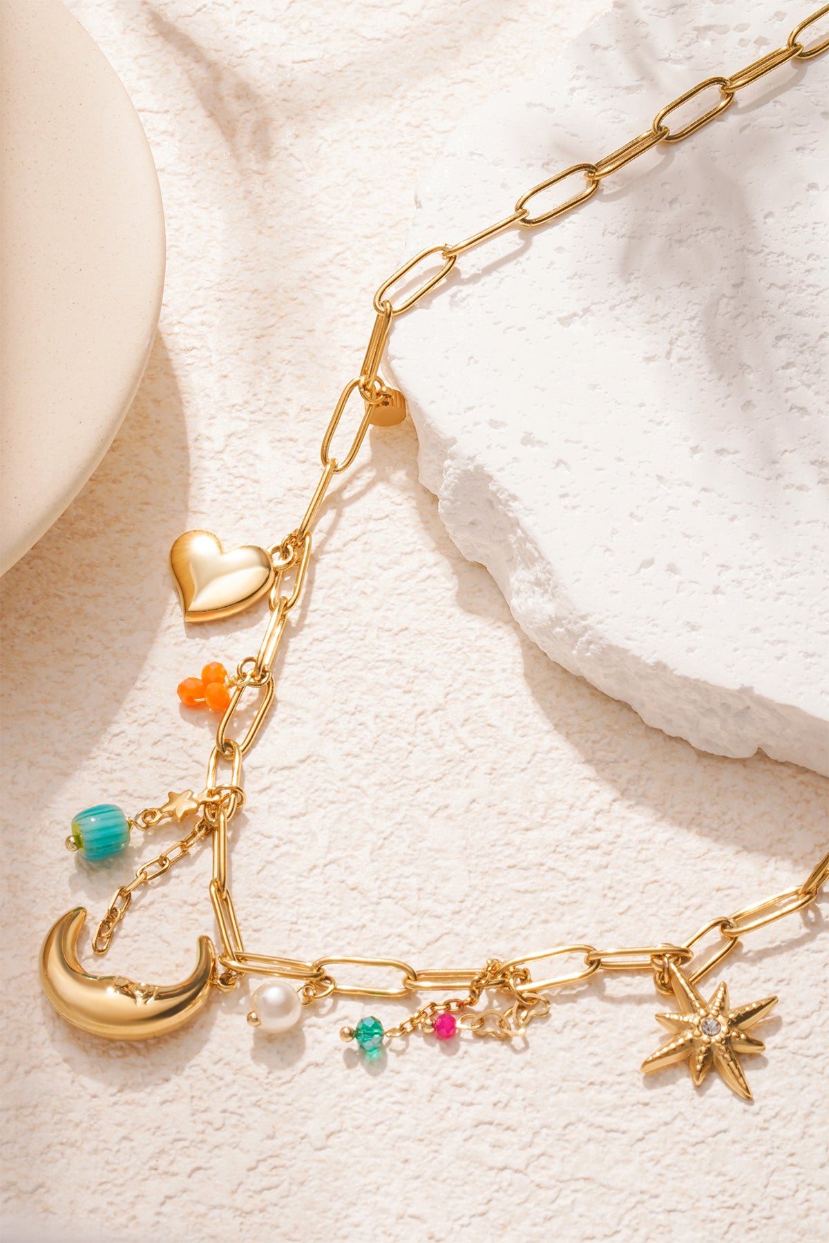 Detailed view of gold link chain necklace with moon, starburst and colourful charms on cream background