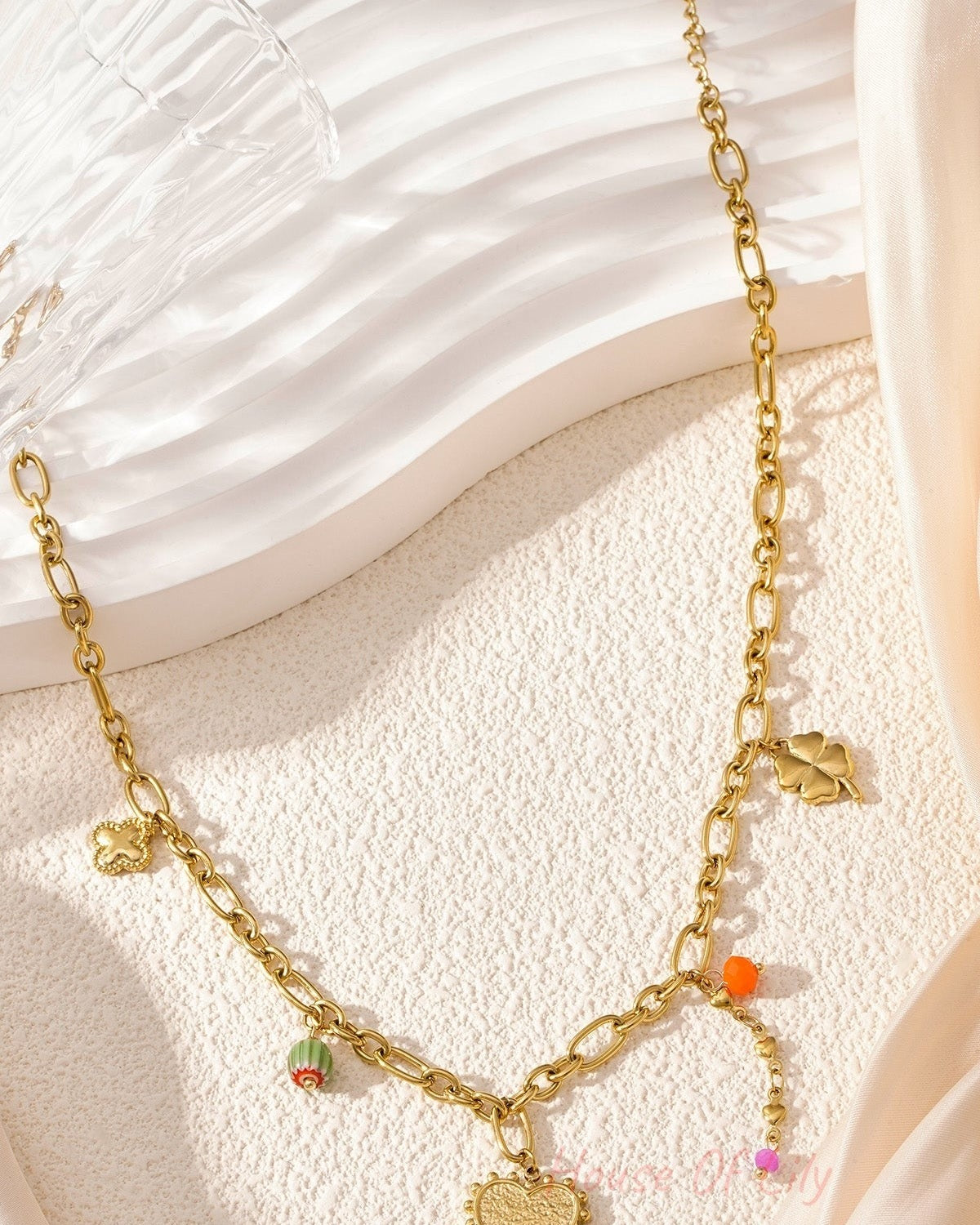 Detailed product shot of golden charm link necklace arranged on a light textured background with a glass, showing the shine of the gold and colourful charms.