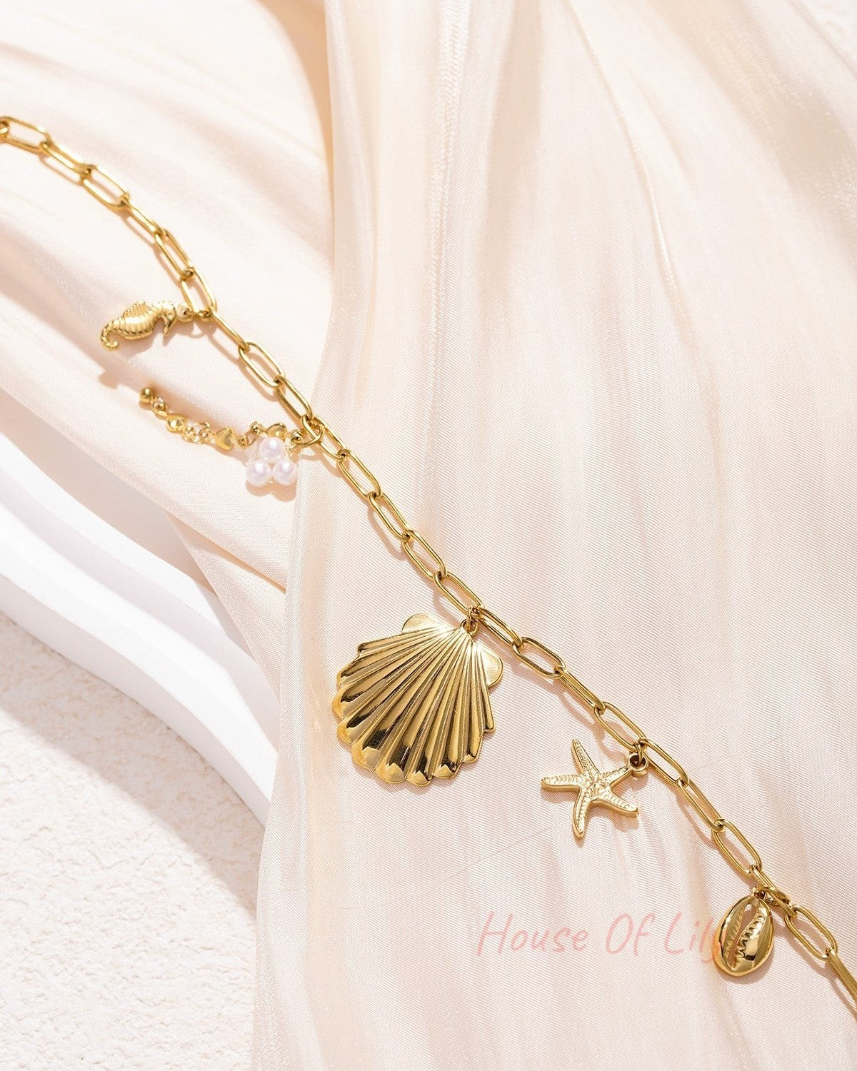 Detailed view of the gold seaside charm necklace with links and charms lying on light-coloured background showing pearl cluster and cowrie shell.