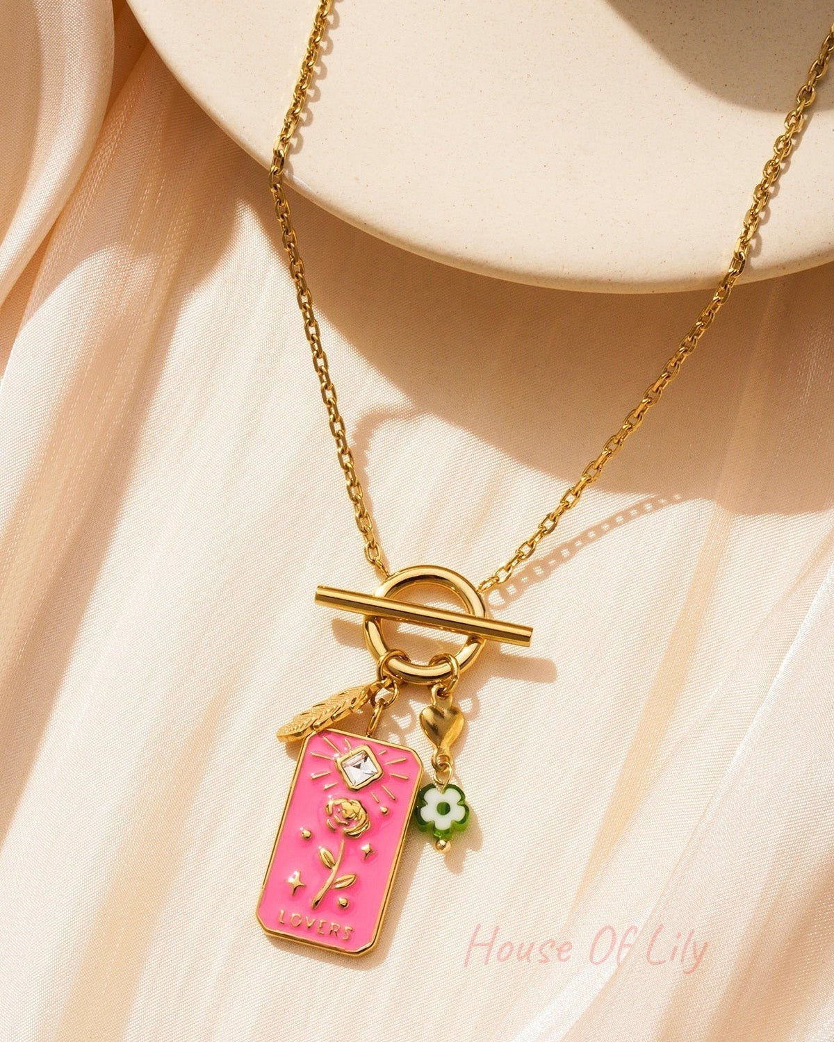 Close-up of the necklace charms with pink floral tag and gold toggle clasp on soft beige fabric background