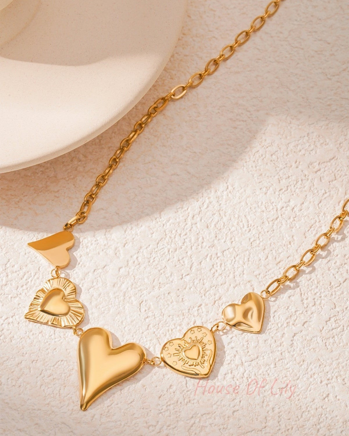 Side angled view of gold multi heart charm necklace on a cream surface showing chain and charm textures