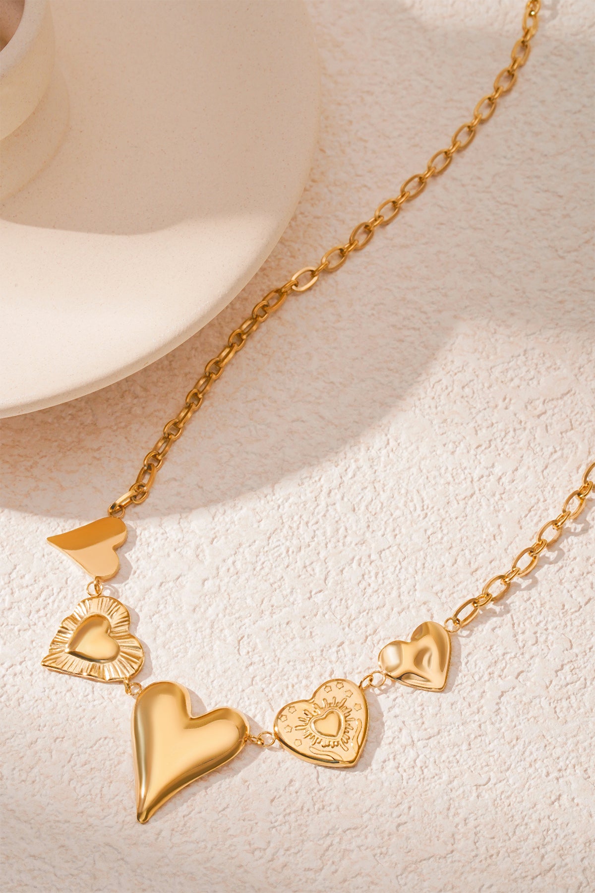 Side angled view of gold multi heart charm necklace on a cream surface showing chain and charm textures