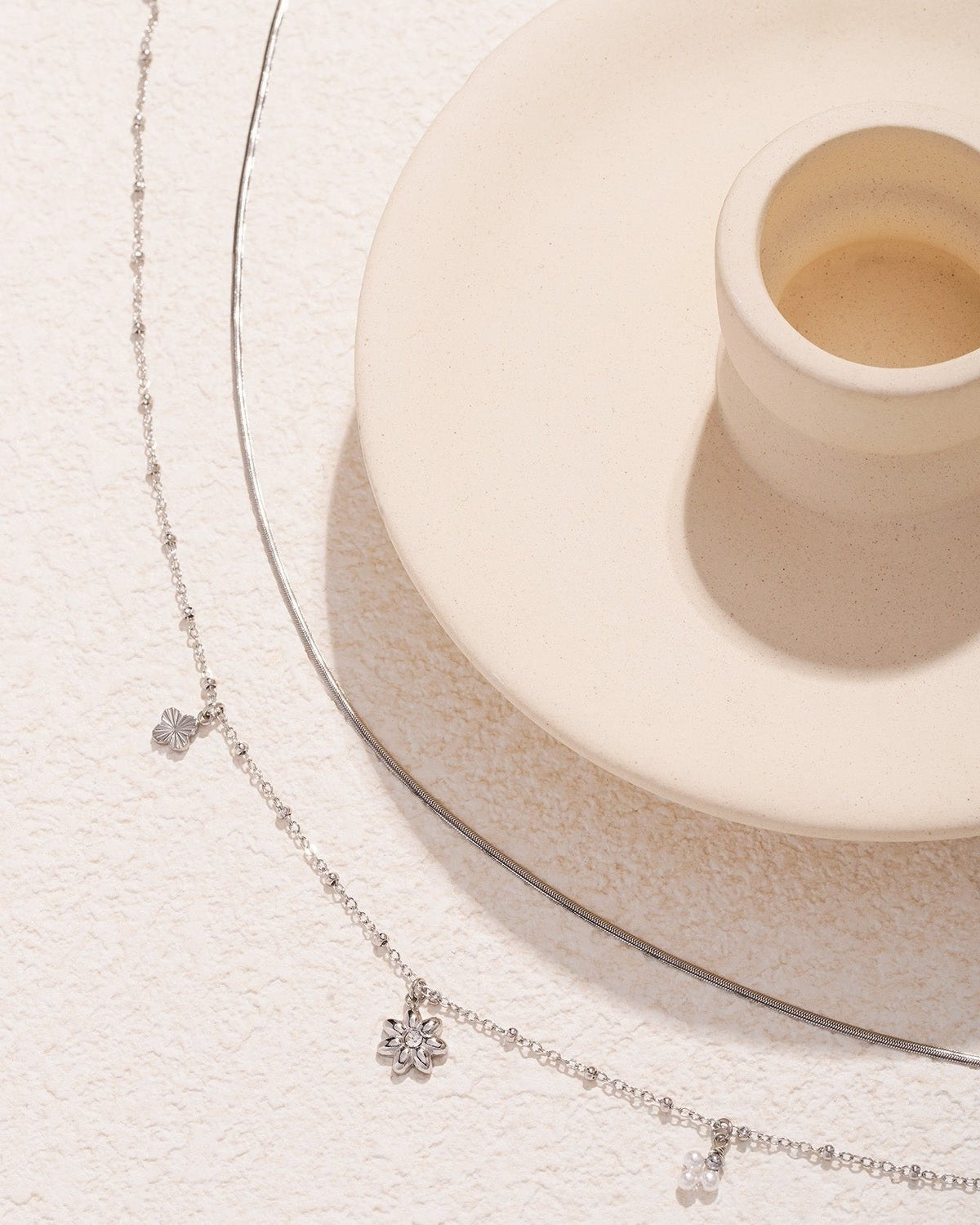 Silver layered necklace with flower and pearl charms detail with beige ceramic cup on textured background