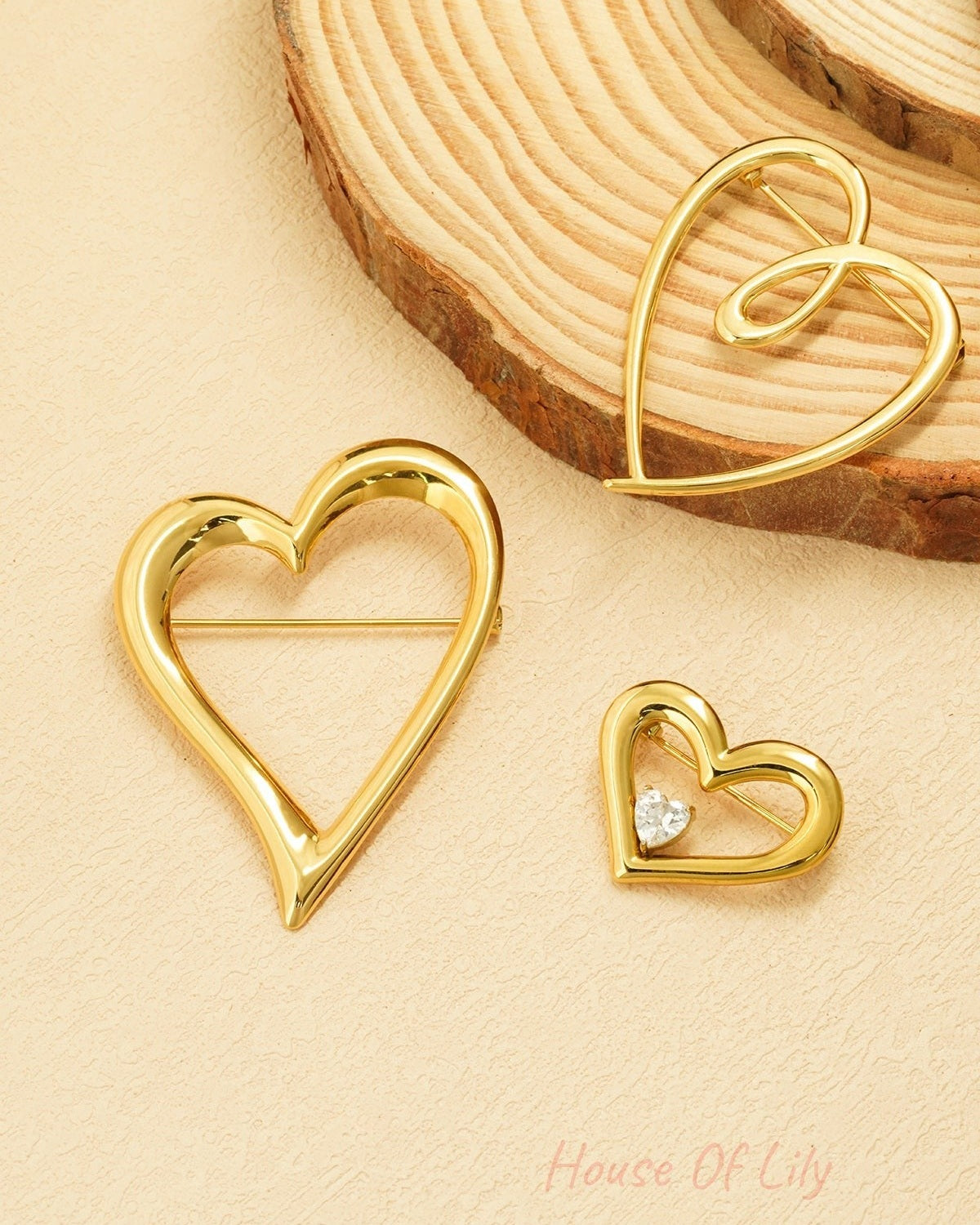 Three gold heart brooches of varying sizes displayed on a natural wood and cream textured surface