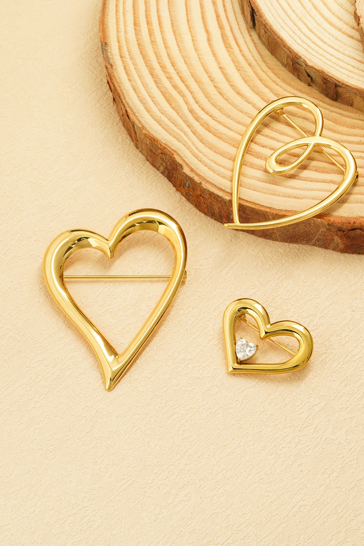 Three gold heart brooches of varying sizes displayed on a natural wood and cream textured surface