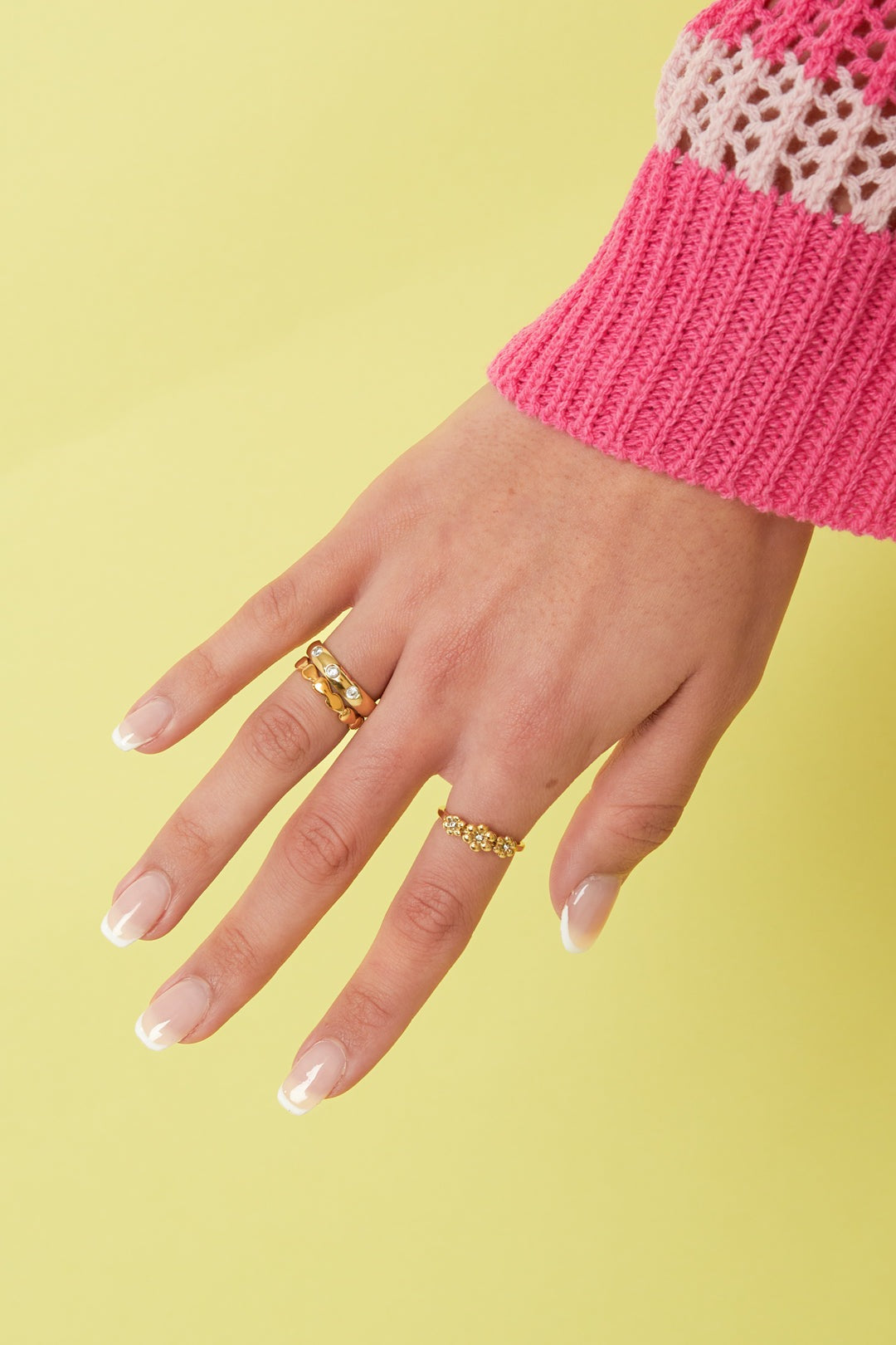 Model hand wearing three gold rings including a gold stone ring, styled with a pink knitted sweater against yellow background