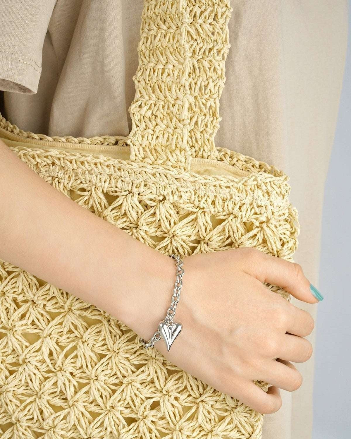 Silver chunky heart charm bracelet worn on wrist holding a woven beige tote bag