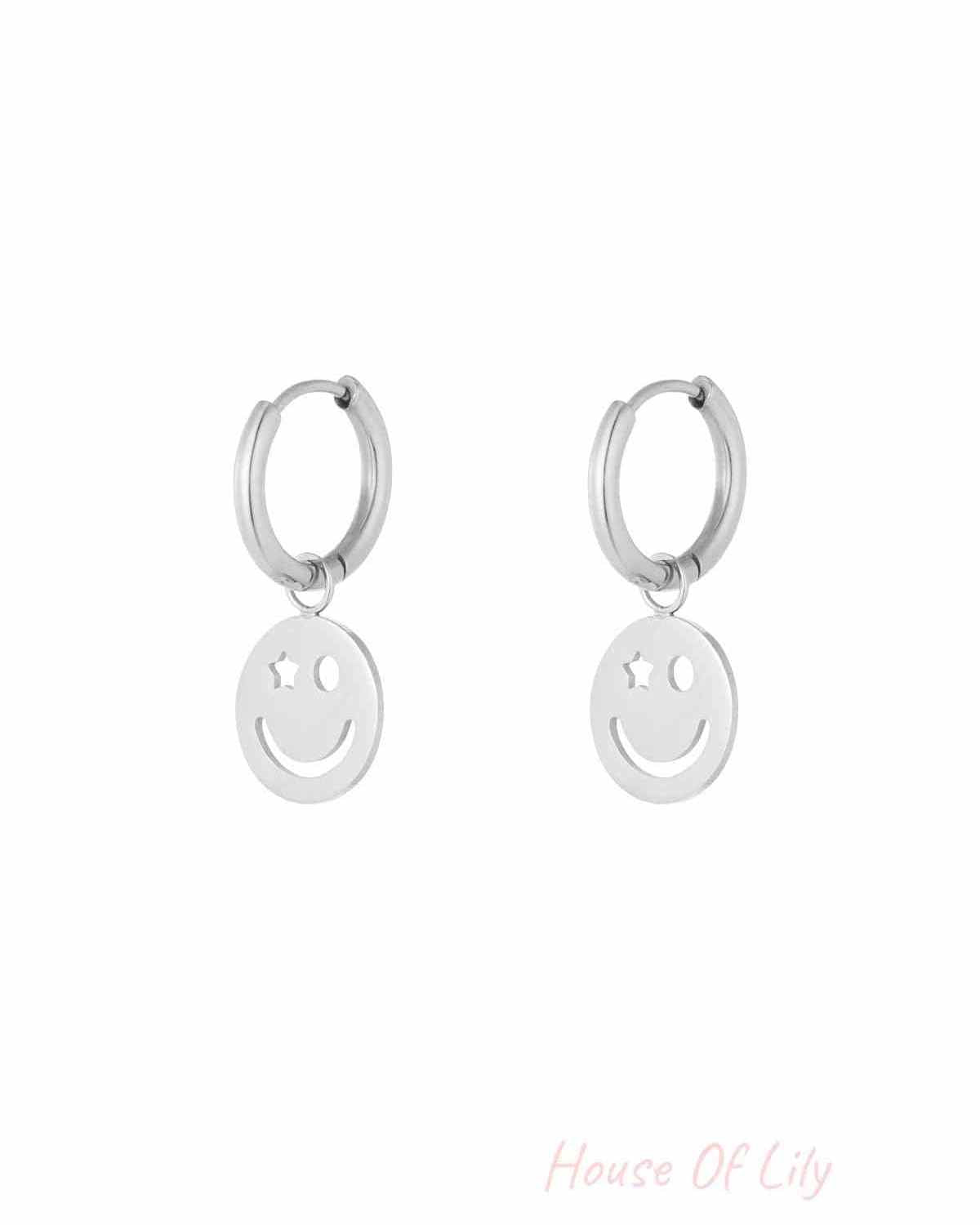 Pair of silver hoop earrings with dangling smiley face charm featuring one star eye and one circular eye on white background