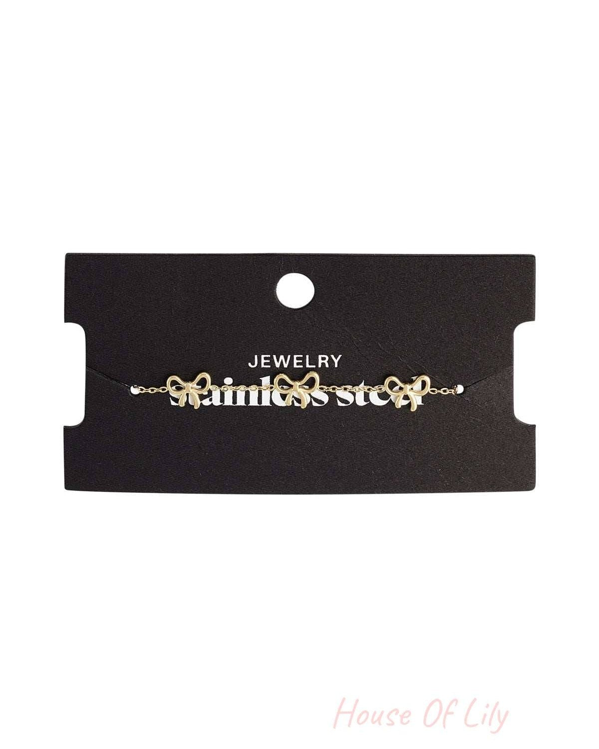 Gold bow charm bracelet displayed on black jewelry card with 'stainless steel' branding