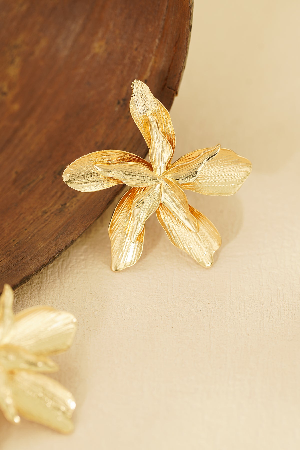 Individual gold flower earring placed near wooden bowl highlighting texture and shine