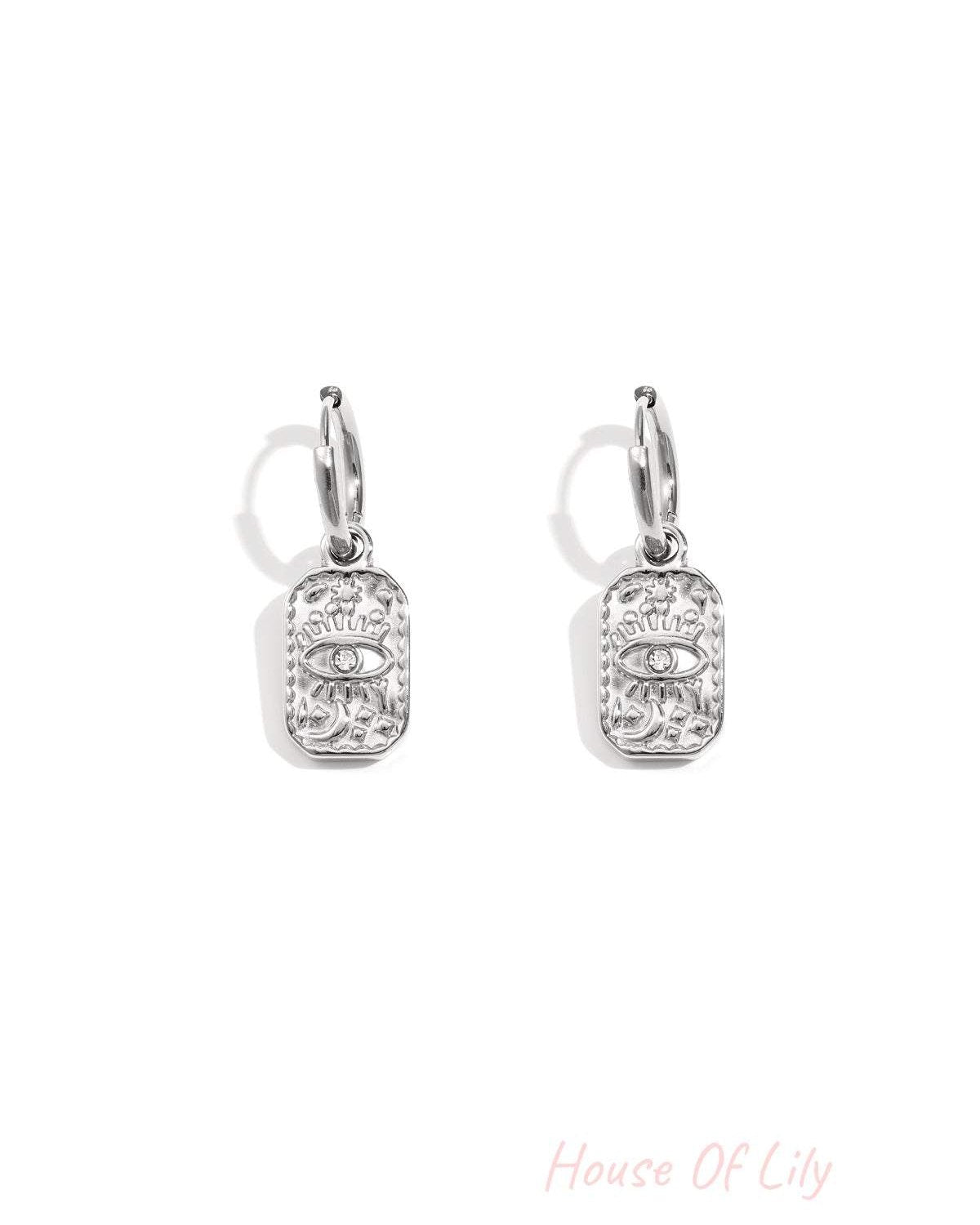 Pair of silver hoop earrings with a rectangular charm embossed with an eye motif and small rhinestone on white background