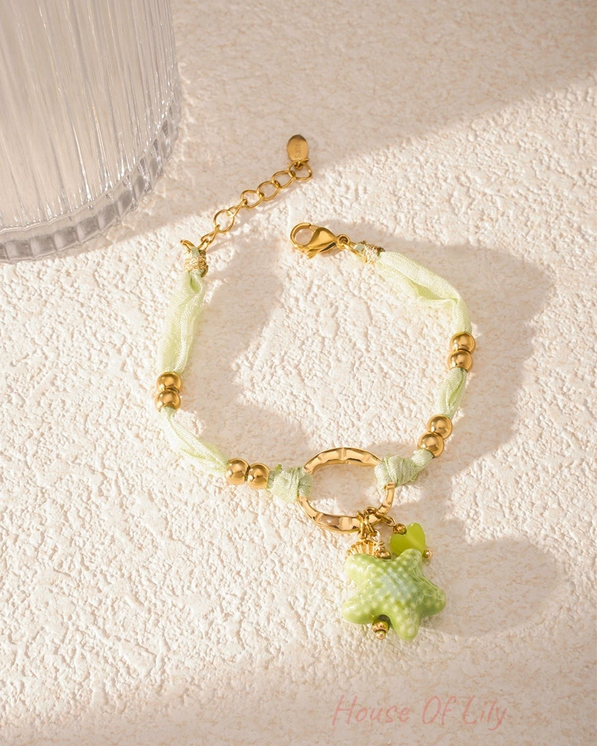 Bracelet lying flat on textured surface with soft sunlight highlighting gold beads and green charms