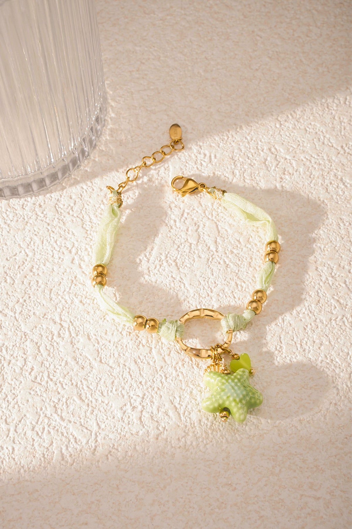 Bracelet lying flat on textured surface with soft sunlight highlighting gold beads and green charms